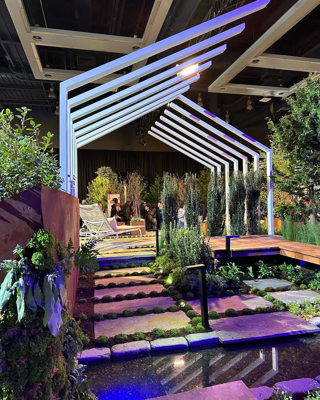 Modern indoor garden with geometric canopy, lush greenery, and stone path. People gather in background, creating a vibrant, eco-friendly atmosphere under ambient lighting.