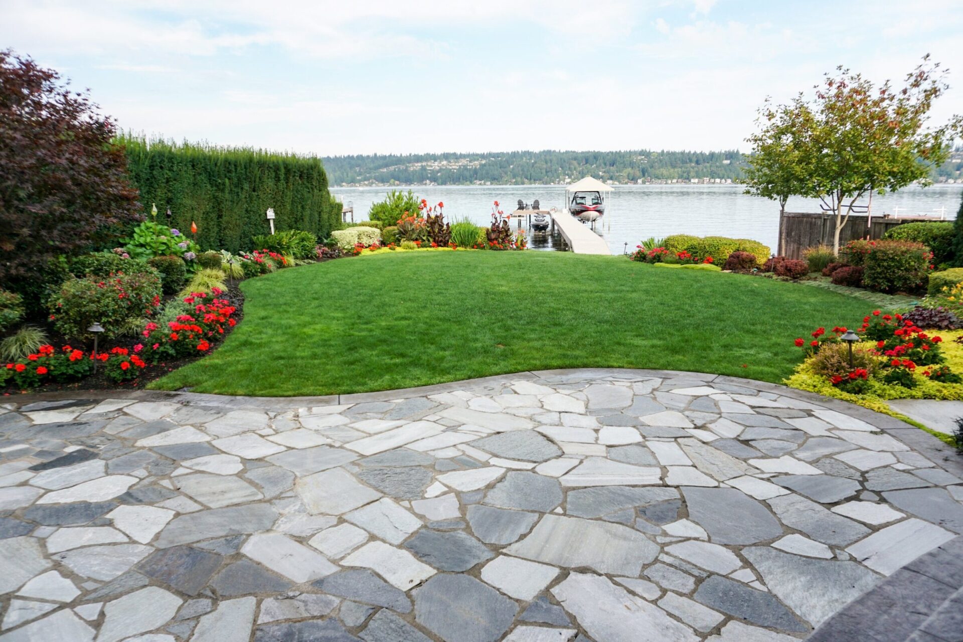 A landscaped garden with vibrant flowers and stone paths overlooking a calm lake, with a small dock and distant forested shoreline.