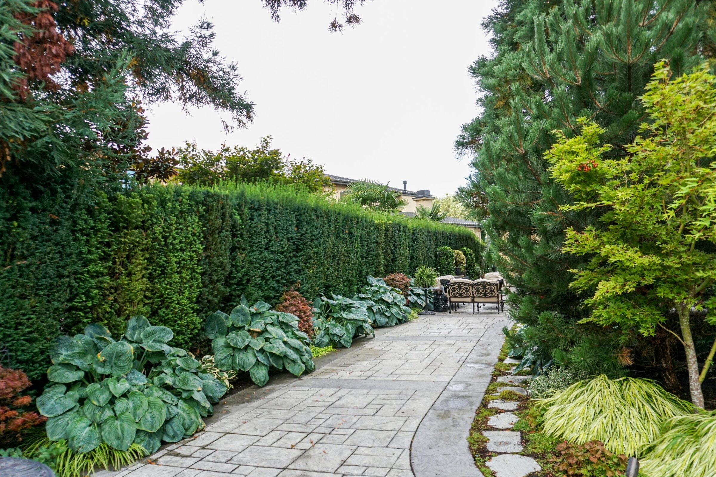 A stone pathway lined with lush greenery leads to a seating area, nestled among tall hedges and trees. No people or landmarks visible.