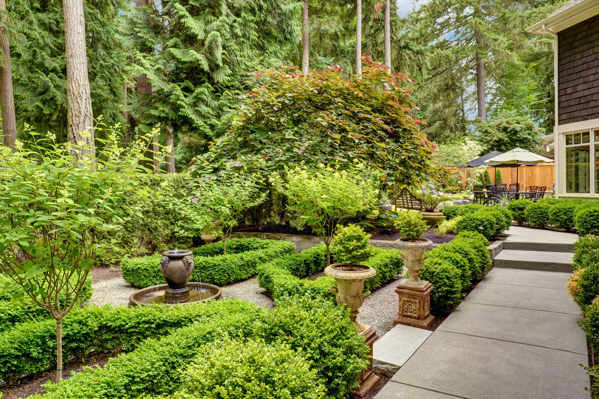 A lush garden with manicured hedges, a fountain, and statues, surrounded by tall trees. Patio furniture is visible in the background.