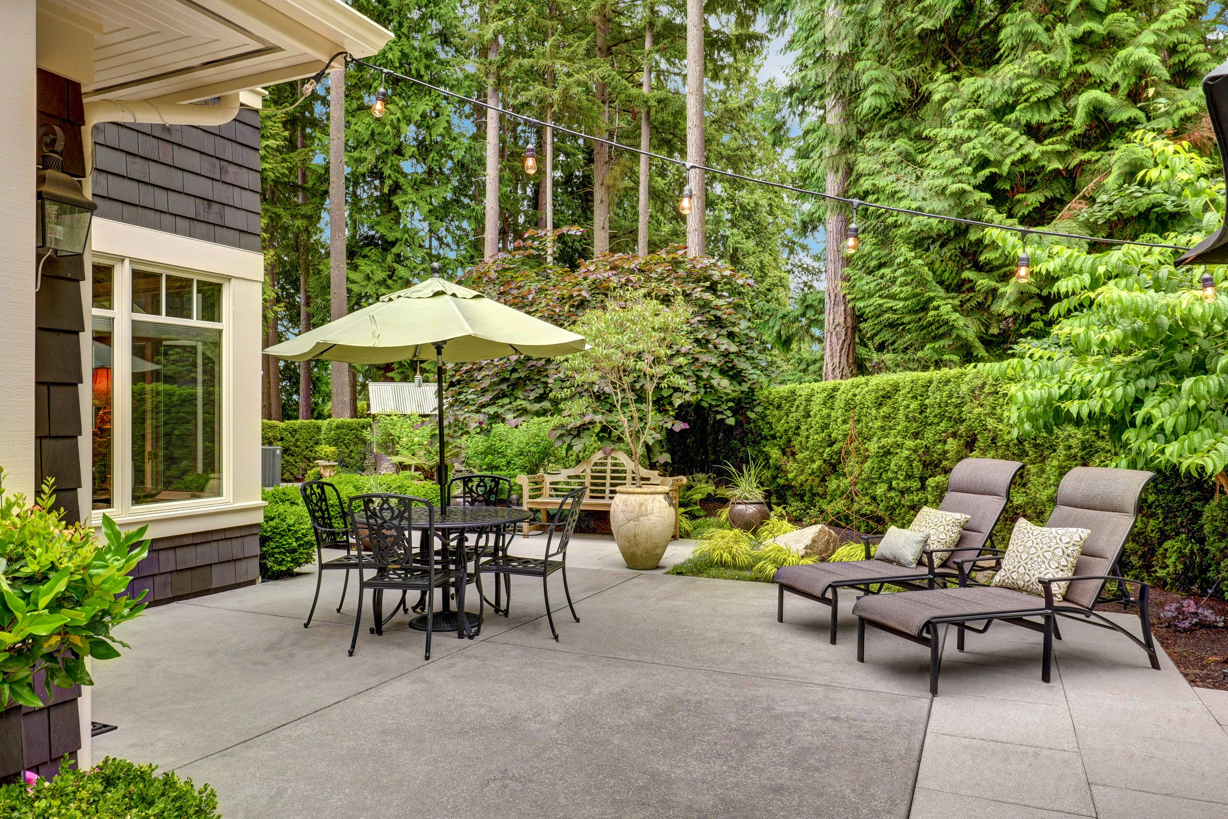 A serene backyard patio features a table with umbrella, lounge chairs, and lush green foliage, creating a tranquil outdoor relaxation spot.