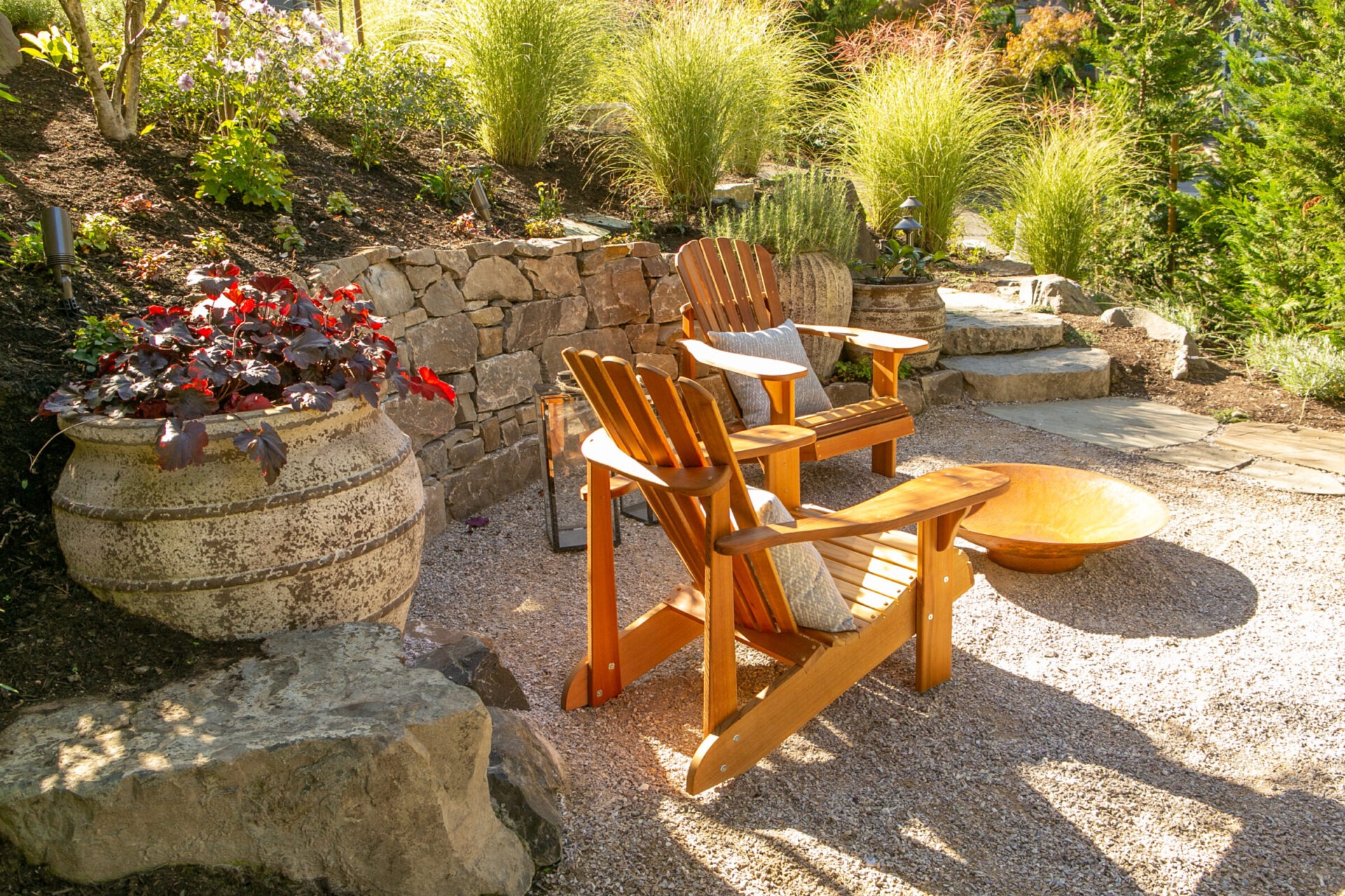 A cozy outdoor area with wooden chairs, fire pit, stone path, and lush greenery, perfect for relaxing in a garden setting.