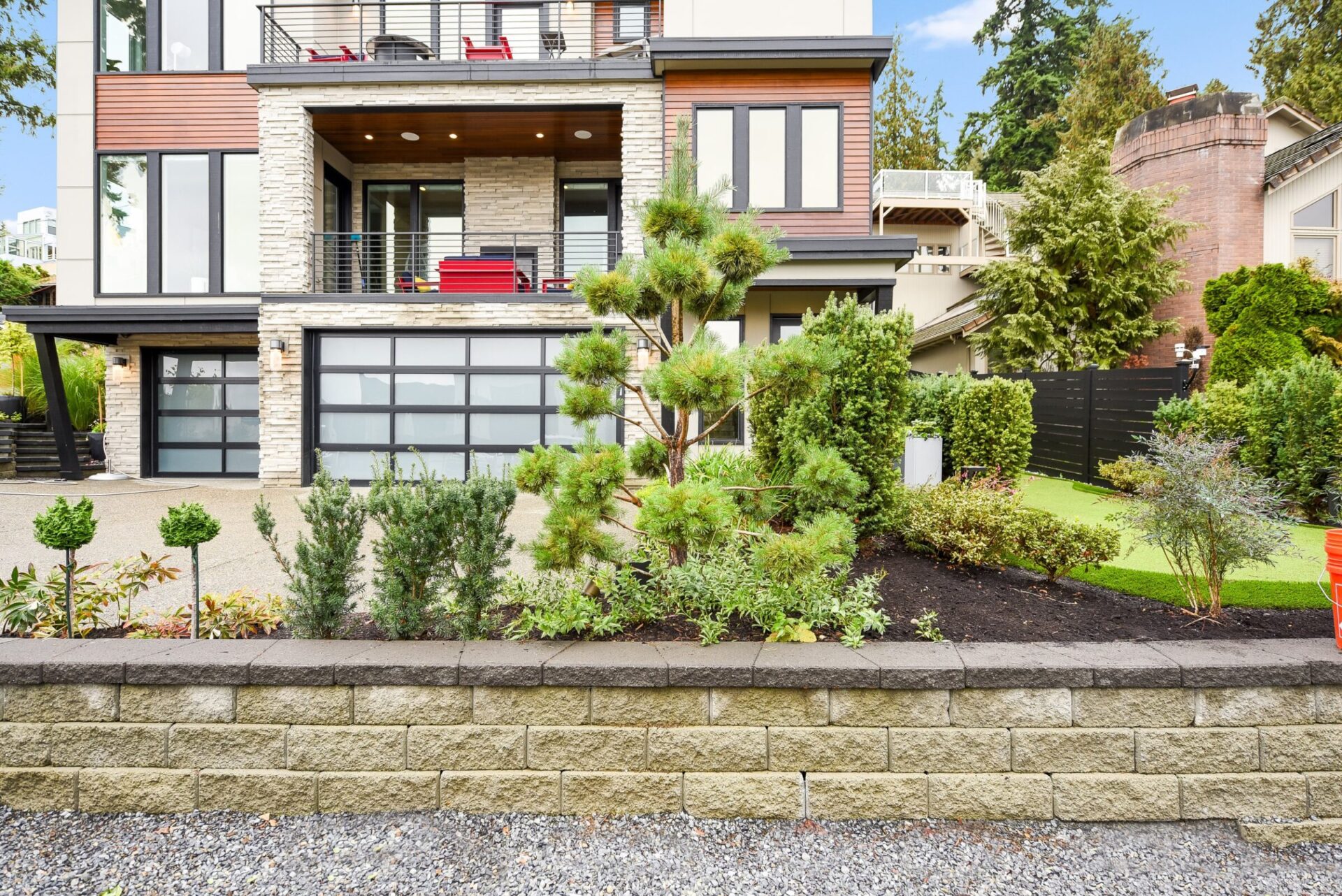 Modern multi-story home with large windows, stone facade, and glass garage door. Manicured garden in front with various trees and shrubs. No landmarks visible.