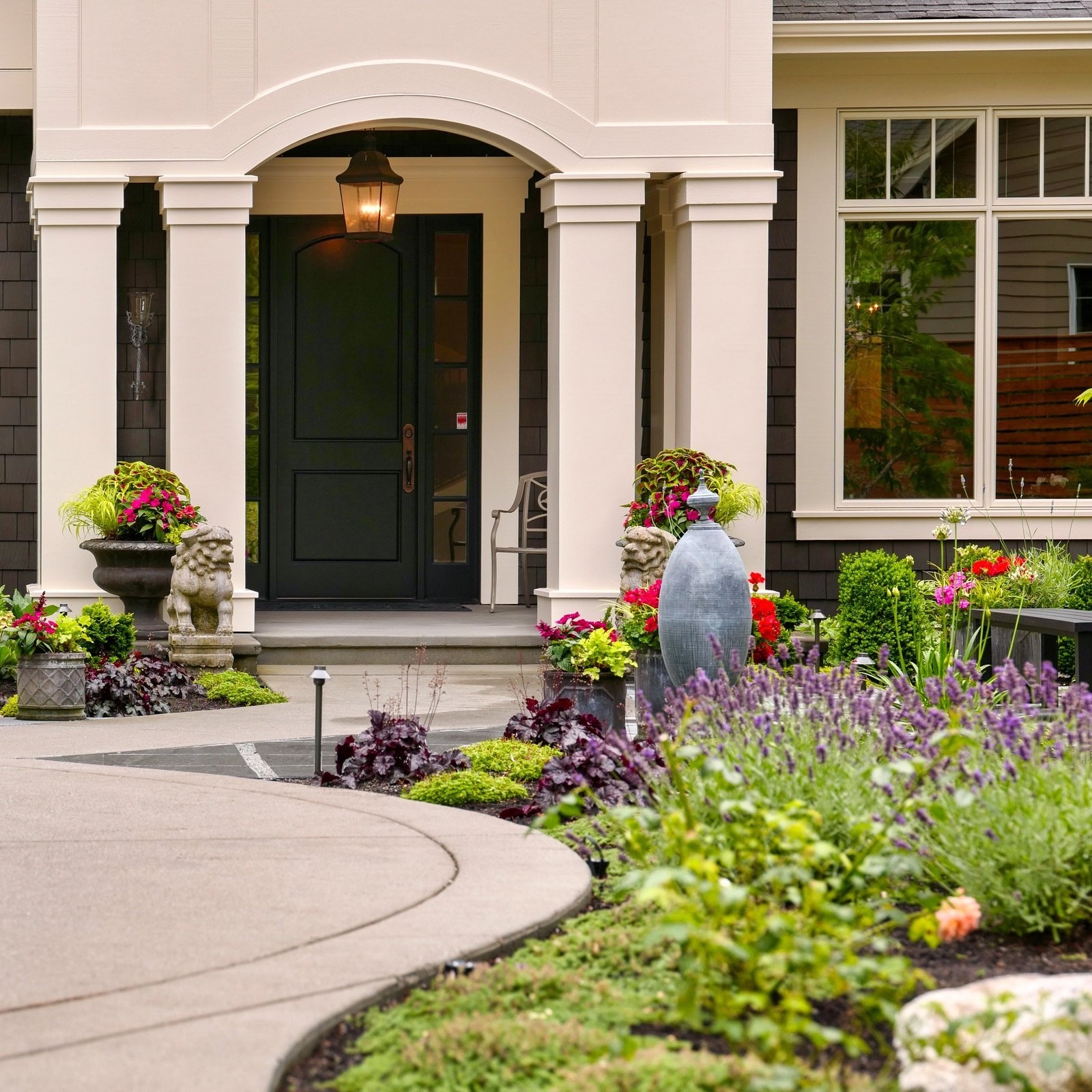 Residential entrance with elegant landscaping, a curved walkway, and decorative plants. Features a black door under an arched porch with column details.