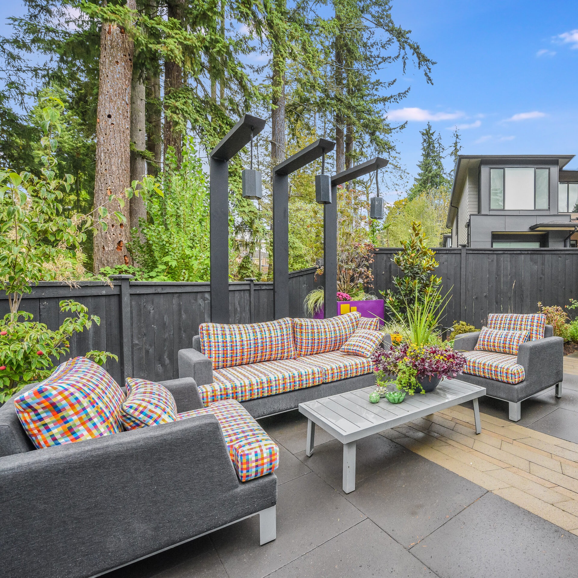 A modern backyard patio features colorful outdoor seating, a coffee table, lush greenery, and tall trees. The space is enclosed by a fence.