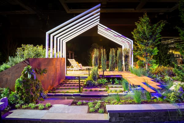 Illuminated modern garden with geometric structure, surrounded by diverse greenery. Central pathway leads to lounge chairs, creating a tranquil, inviting atmosphere.