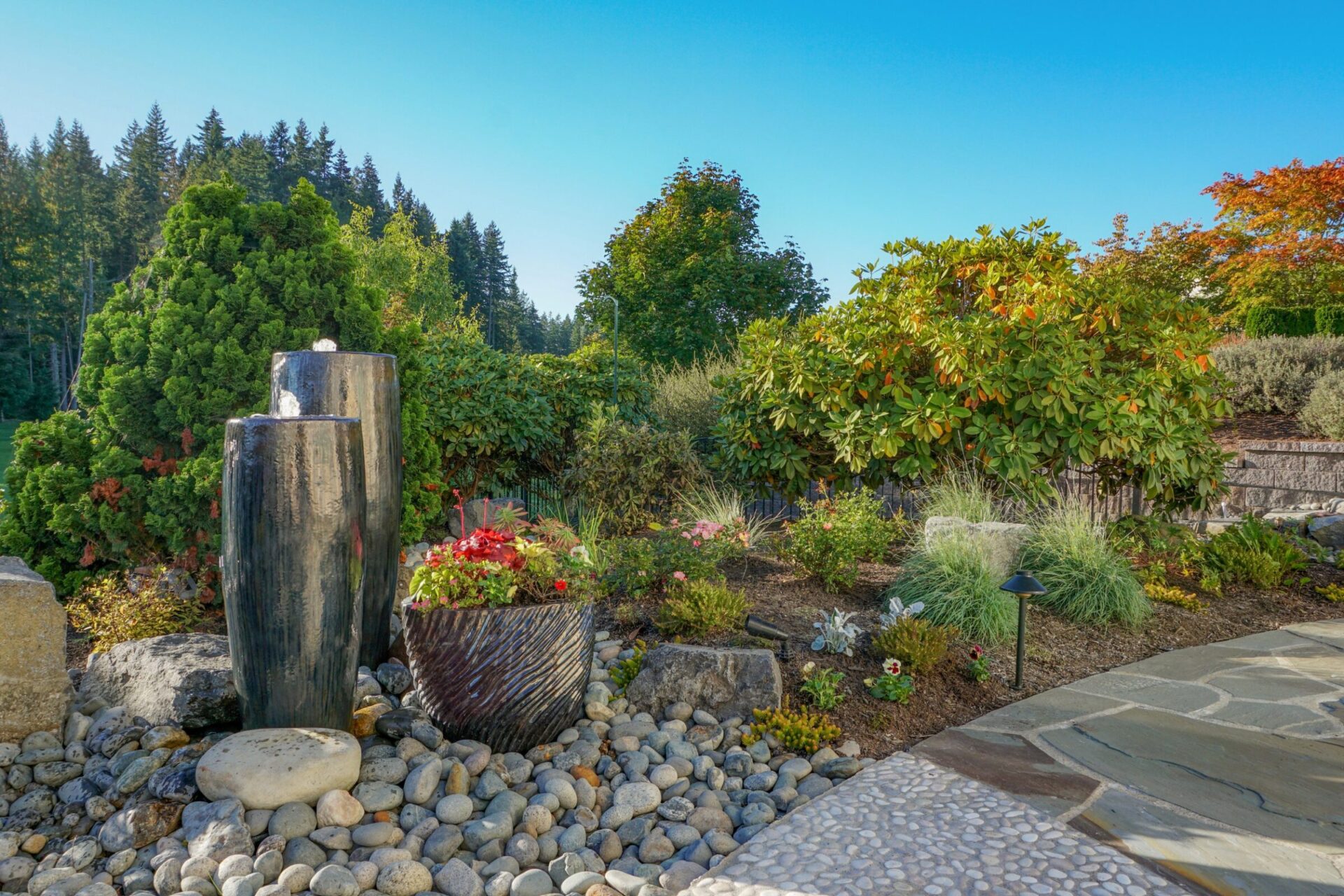 A serene garden scene with stone paths, lush greenery, and a water feature. Decorative plants and rocks enhance the tranquil atmosphere under a clear sky.