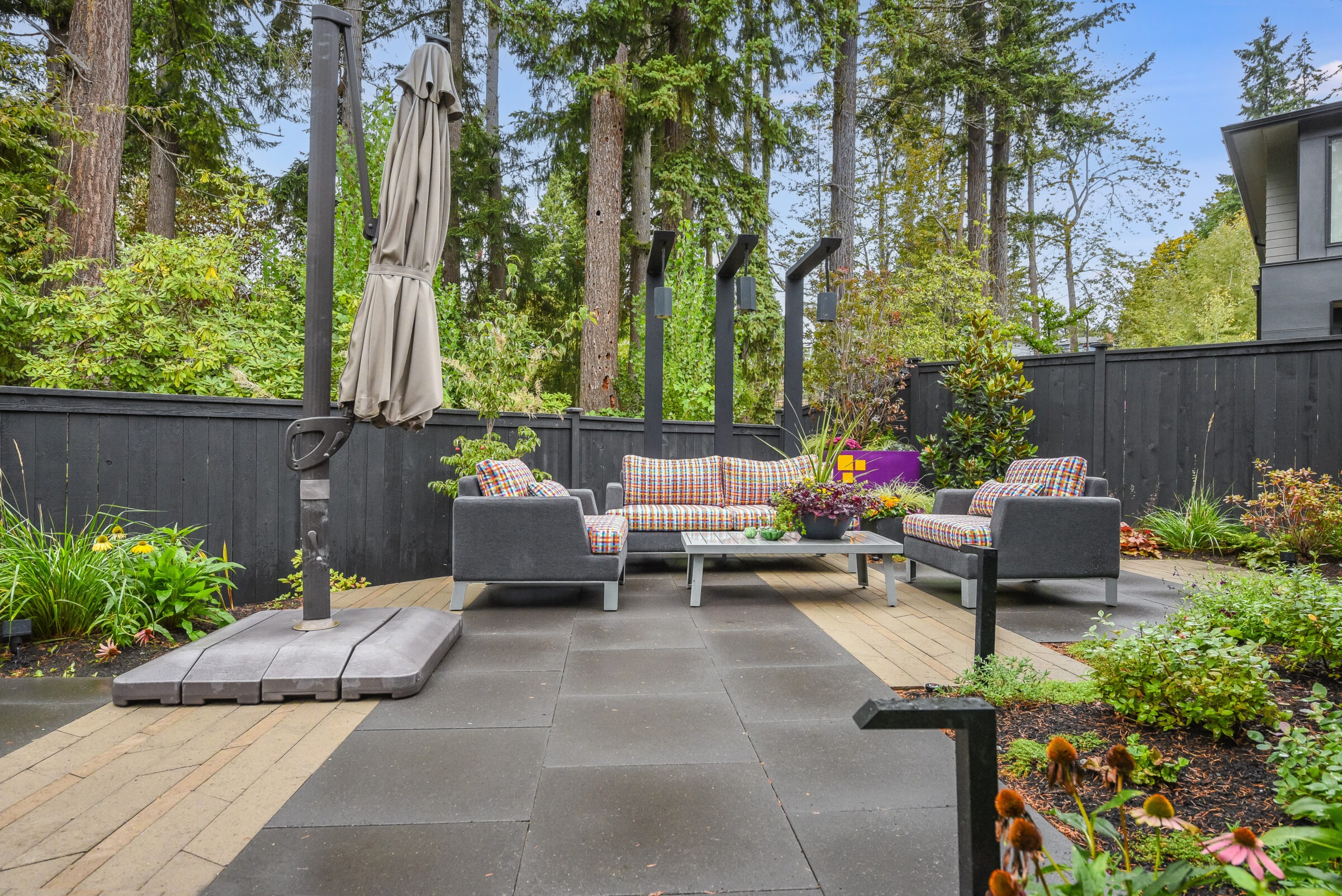 Modern outdoor patio with cushioned seating, surrounded by lush greenery and tall trees. Features a large umbrella and sleek flooring design.