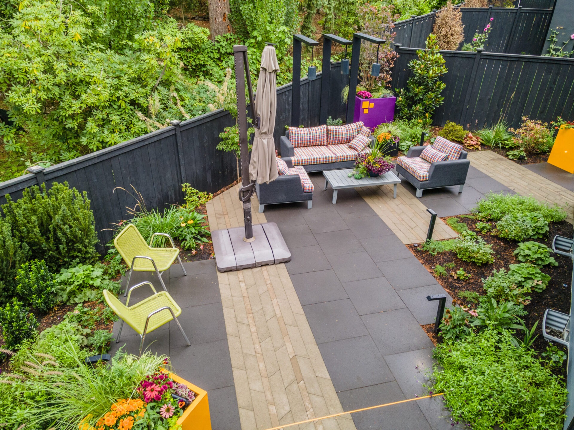 A modern outdoor patio with chairs, couches, umbrellas, and vibrant planters surrounded by lush greenery, enclosed by a dark fence. No people visible.