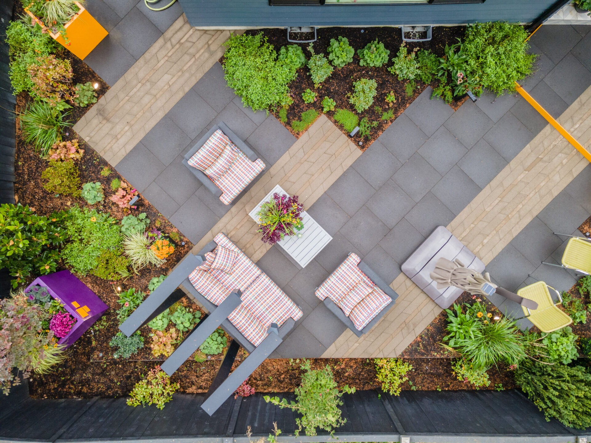 Aerial view of a garden terrace with seating, plants, and patterned pathways. Furniture includes striped cushions and a central coffee table.