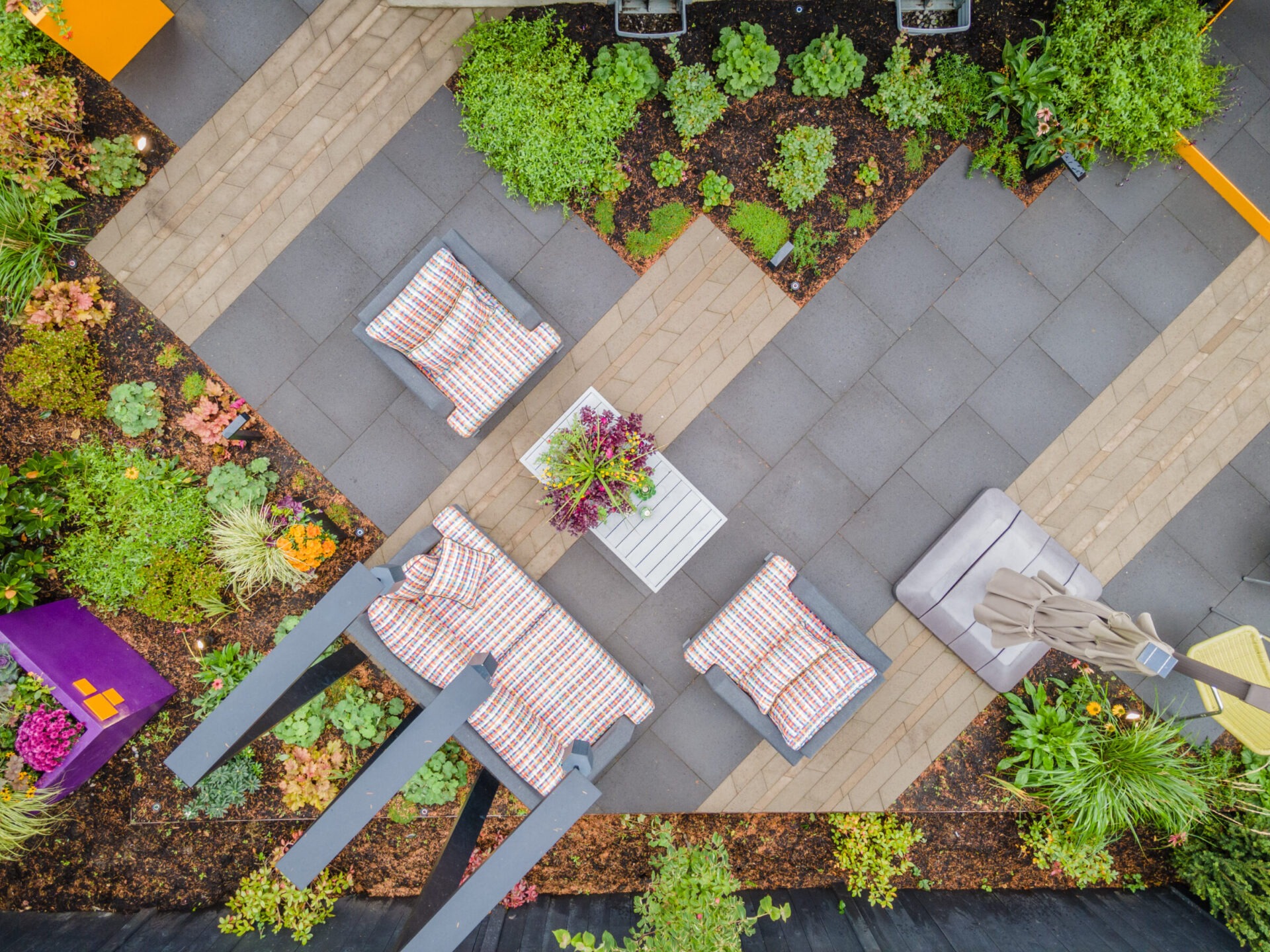 Modern garden with geometric paving, cozy seating, colorful cushions, and lush greenery. A small table with plants provides a focal point.