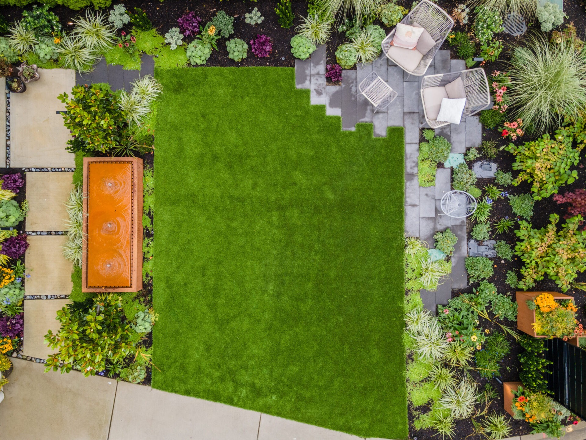 Aerial view of a landscaped garden with a grass lawn, seating area, paved pathway, water feature, and various plants and flowers.