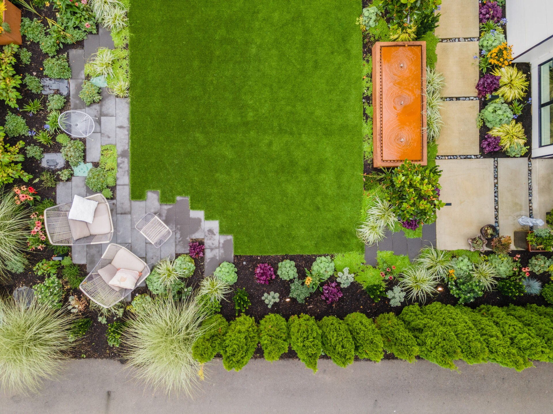 Aerial view of a landscaped garden featuring a green lawn, patio with chairs, pathway, and various plants and shrubs arranged neatly.