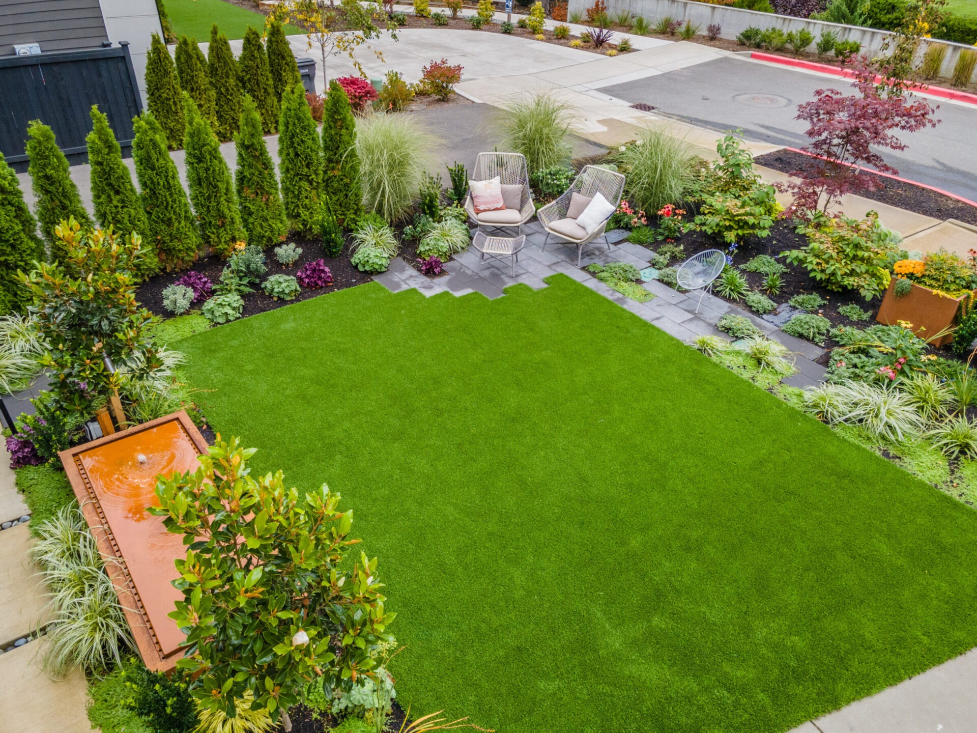 A neatly landscaped backyard features a lush lawn, diverse plants, a fountain, and chairs on a patio, surrounded by modern residential architecture.