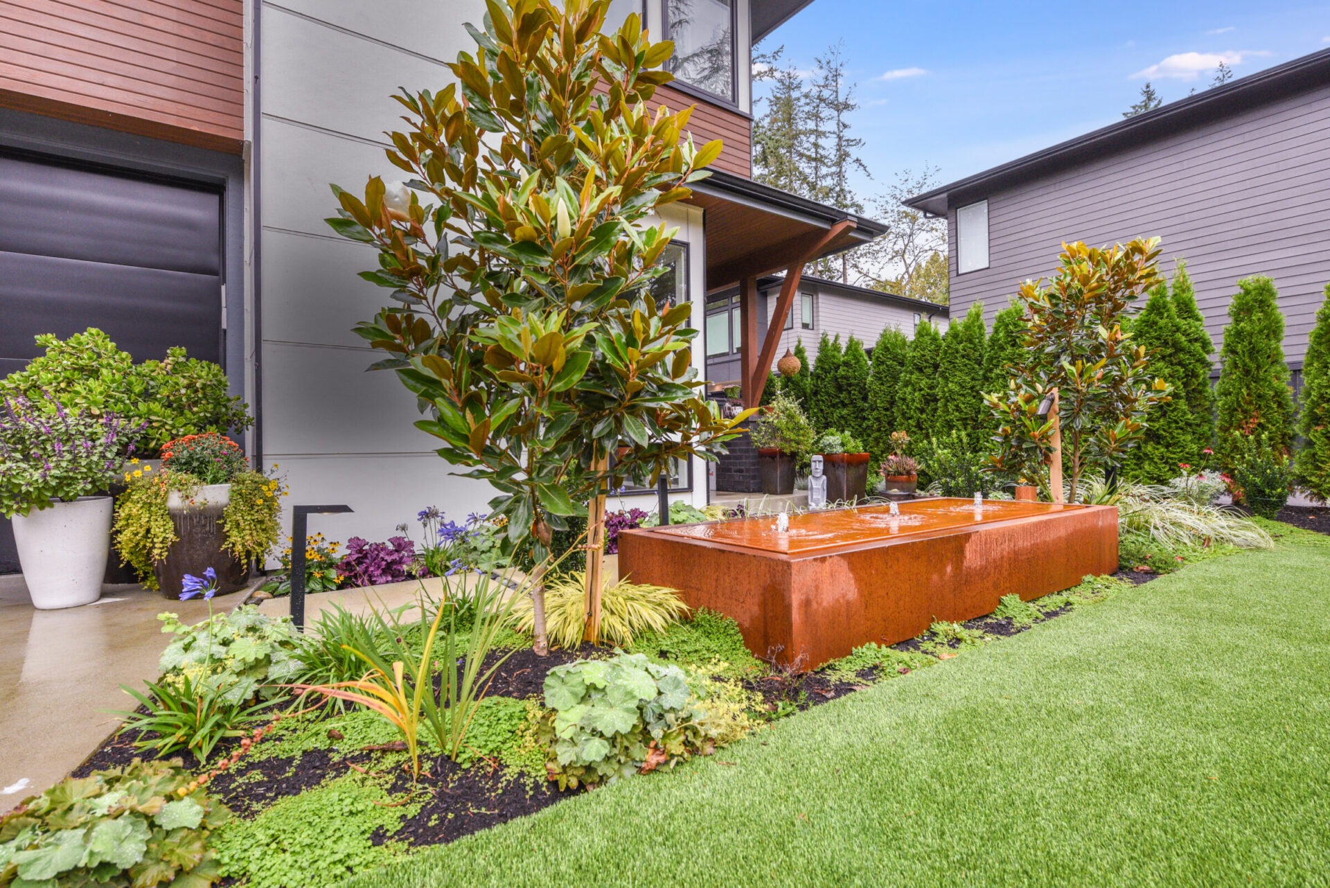 Modern house exterior with a lush garden, featuring manicured grass, various plants, and a decorative metal water feature in a residential neighborhood.