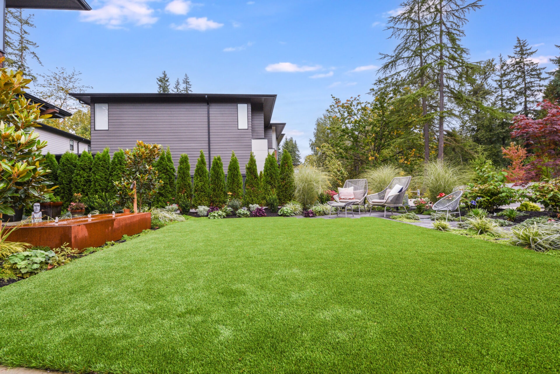 A lush garden with a modern house, featuring manicured grass, trees, planters, and patio chairs under a clear, blue sky.