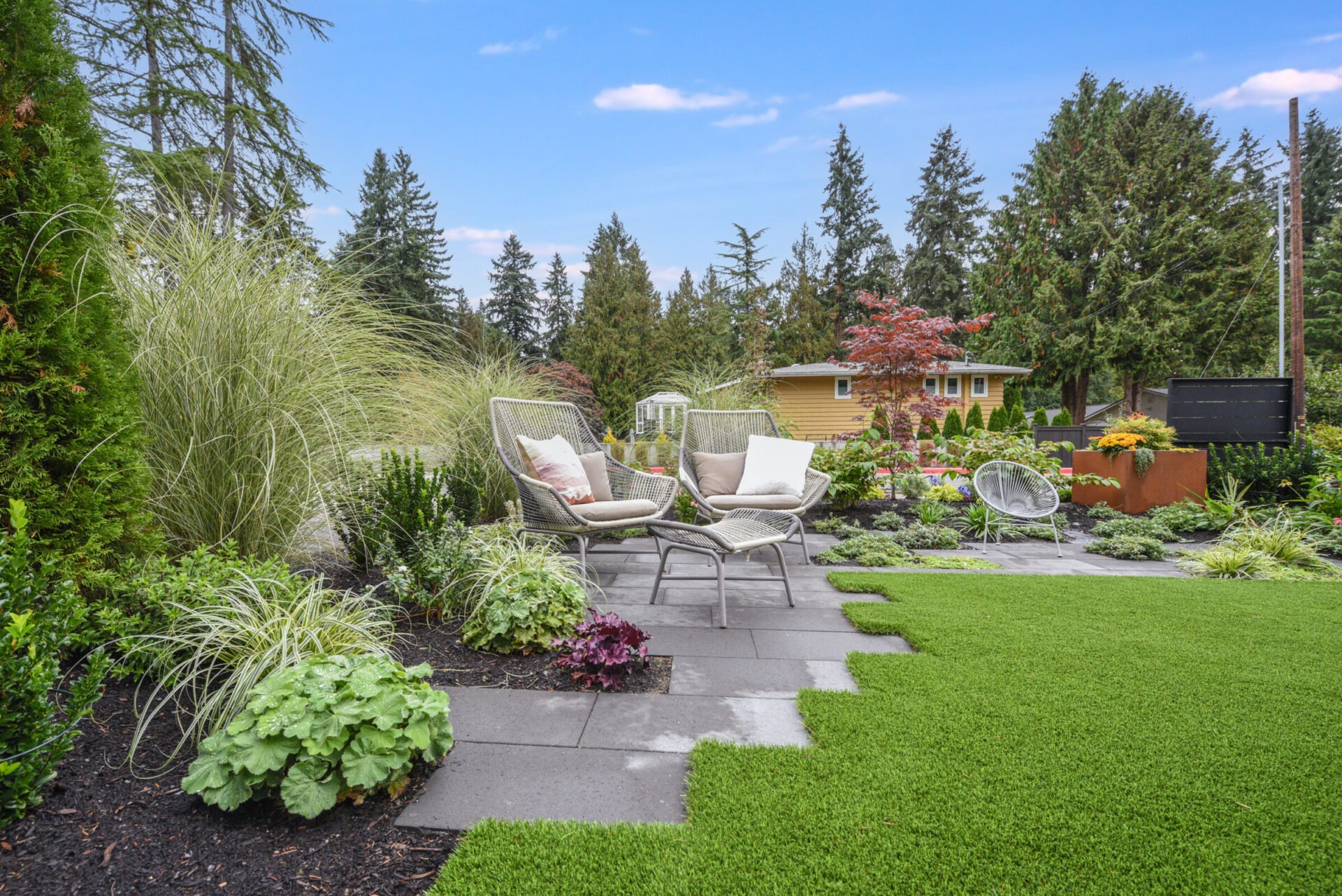 Lush garden with patio seating, surrounded by diverse plants and trees. Cozy outdoor space under a clear blue sky, ideal for relaxation.