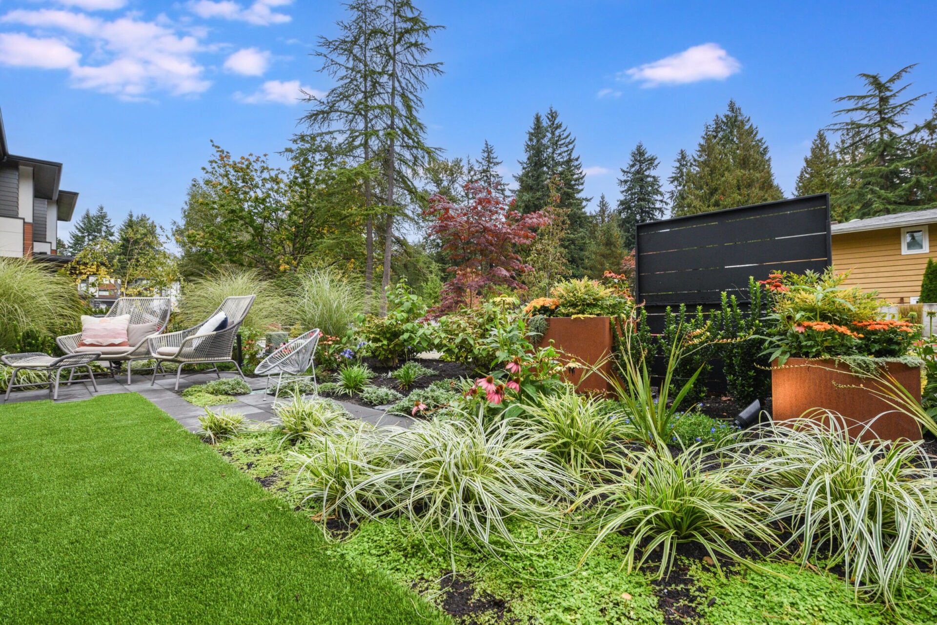 A landscaped backyard features lush greenery, lawn chairs, and vibrant flowers under a clear blue sky. Trees surround the tranquil, modern garden setting.