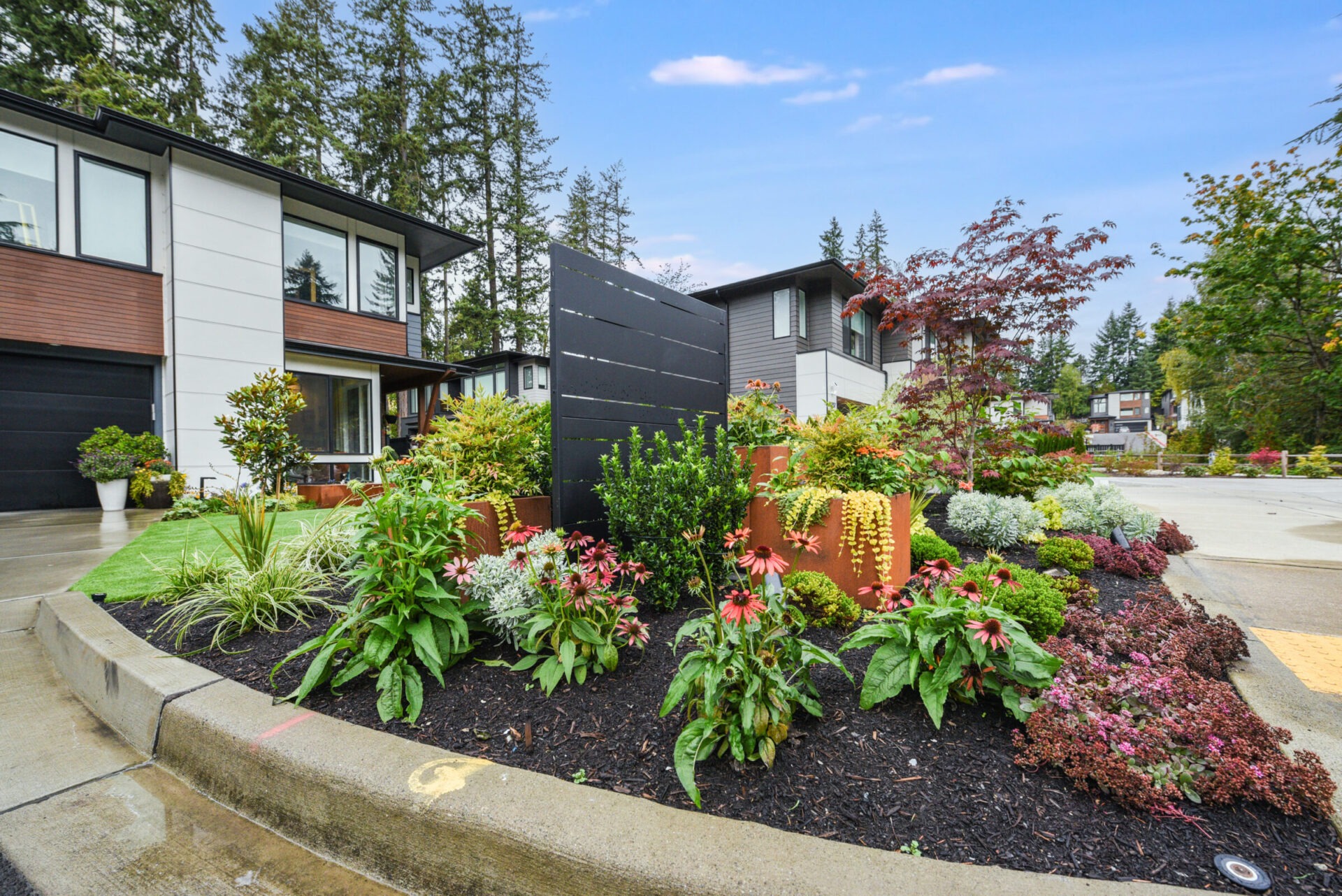 Modern homes with landscaped gardens, vibrant flowers, and trees line a suburban street under a clear sky. No recognizable landmarks or historical buildings visible.