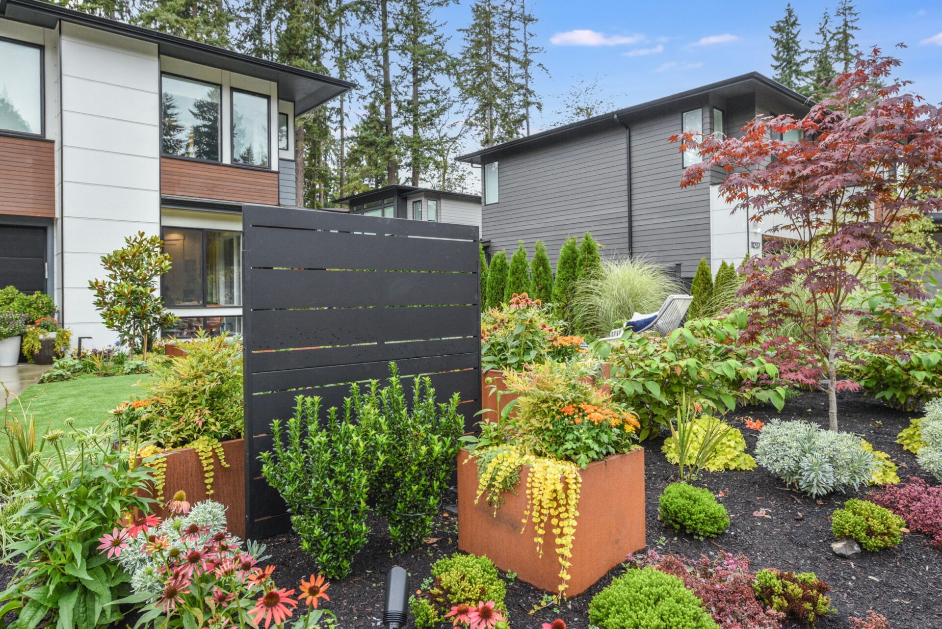 Modern houses with vibrant gardens featuring diverse plants and shrubs. The setting is peaceful, surrounded by tall trees, with no landmarks visible.