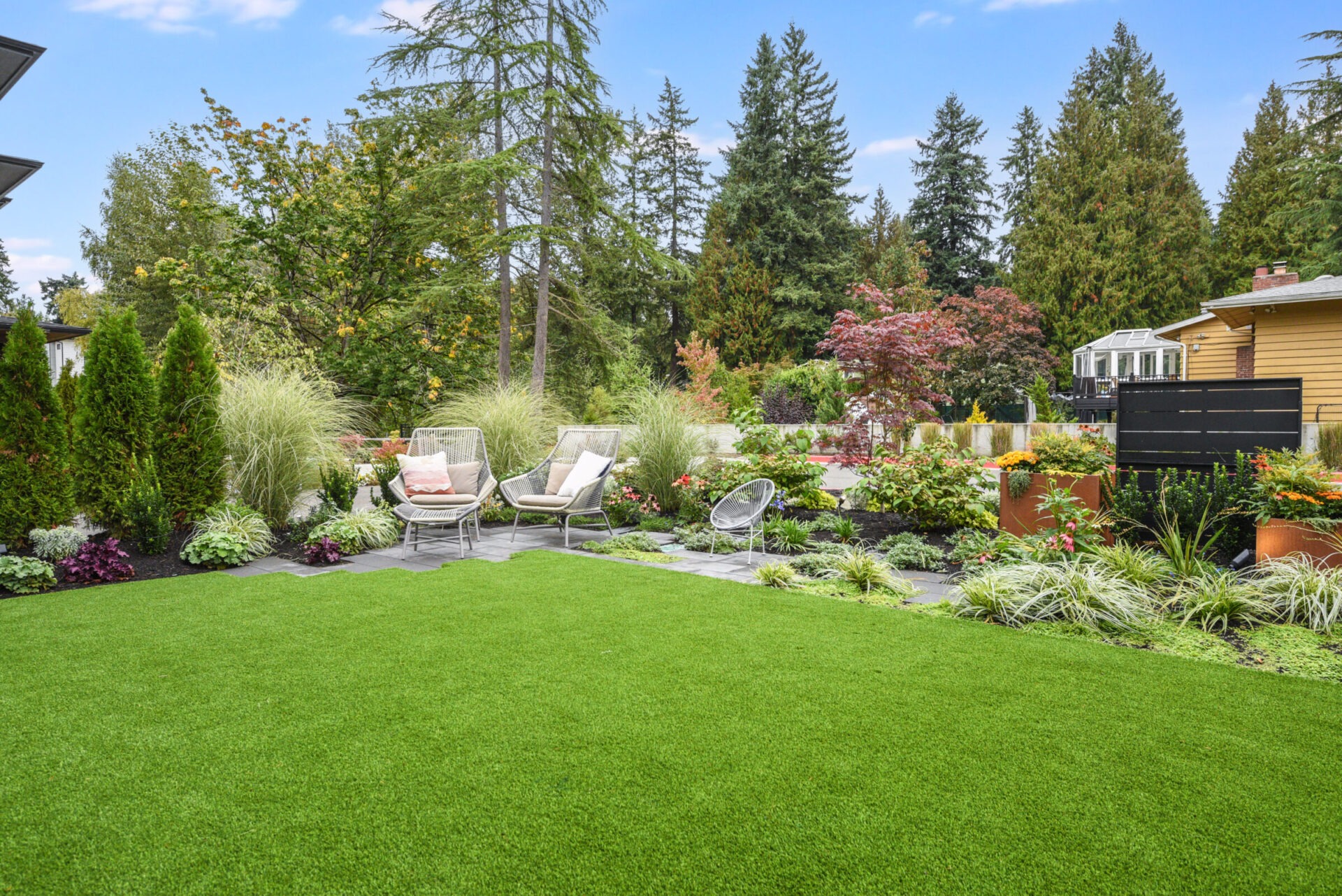 A lush garden with green lawn, surrounded by trees and plants. Two lounge chairs are set on a stone patio under a clear sky.