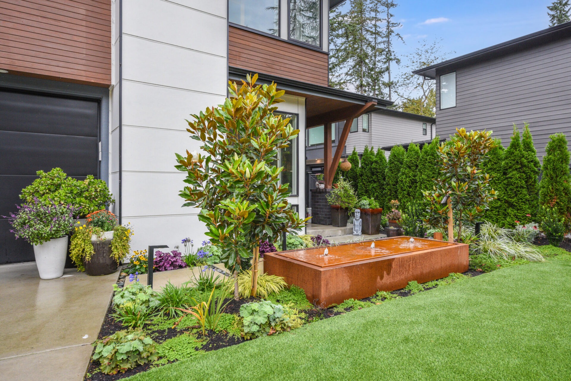 Modern house with a well-maintained garden, featuring a rectangular water fountain, lush greenery, potted plants, and manicured lawn. No people visible.