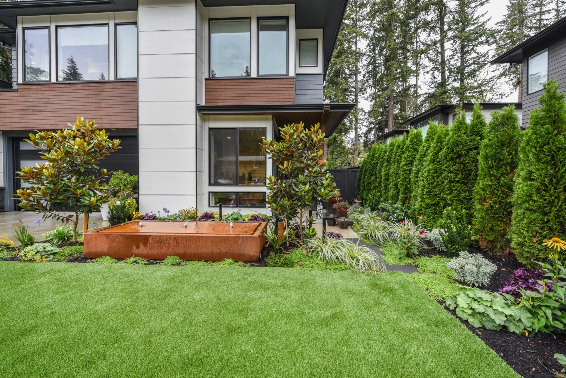 Modern house with manicured garden featuring lush greenery, a small patio, and conifer trees. Large windows and wooden accents emphasize contemporary design.