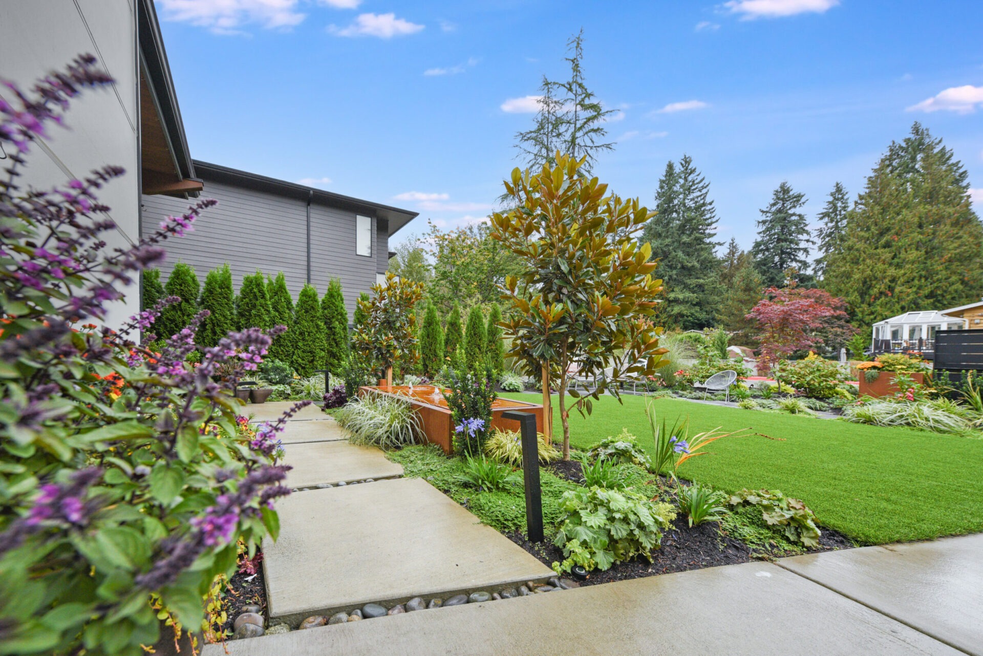 Modern house with landscaped garden, vibrant flowers, and a neat pathway. Surrounded by tall trees under a clear blue sky. No visible landmarks.