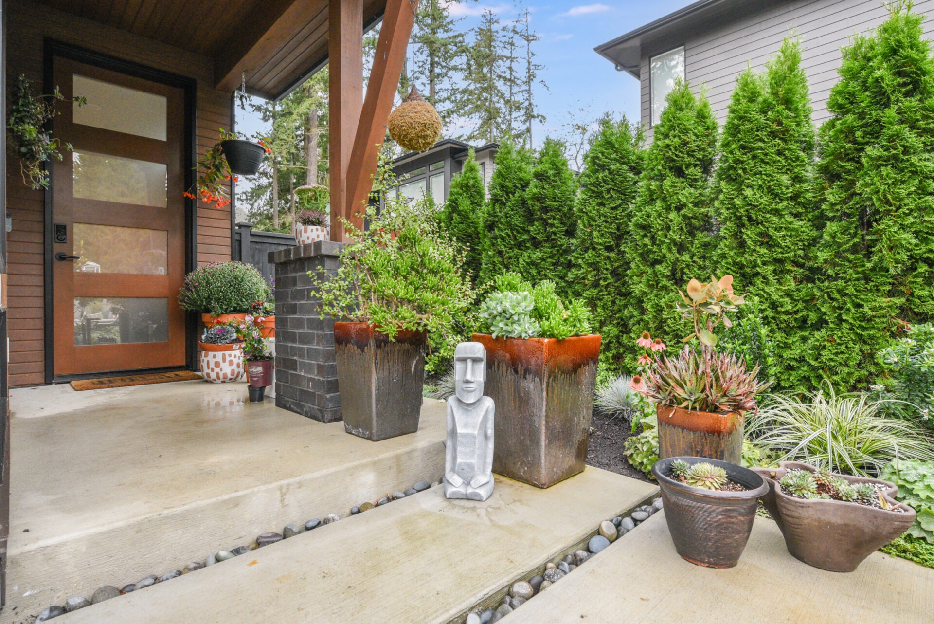 A modern house entrance features lush green plants, a stone statue, and potted flowers, creating a welcoming and serene atmosphere.