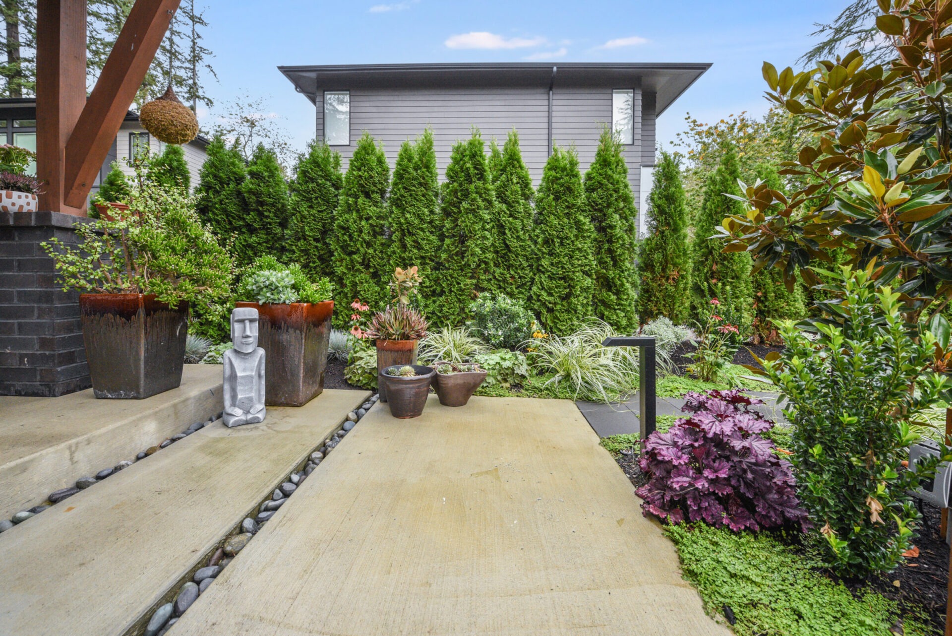 A serene backyard features lush greenery, neatly arranged plants, modern stone path, and a small statue. Privacy hedge frames the light gray house.