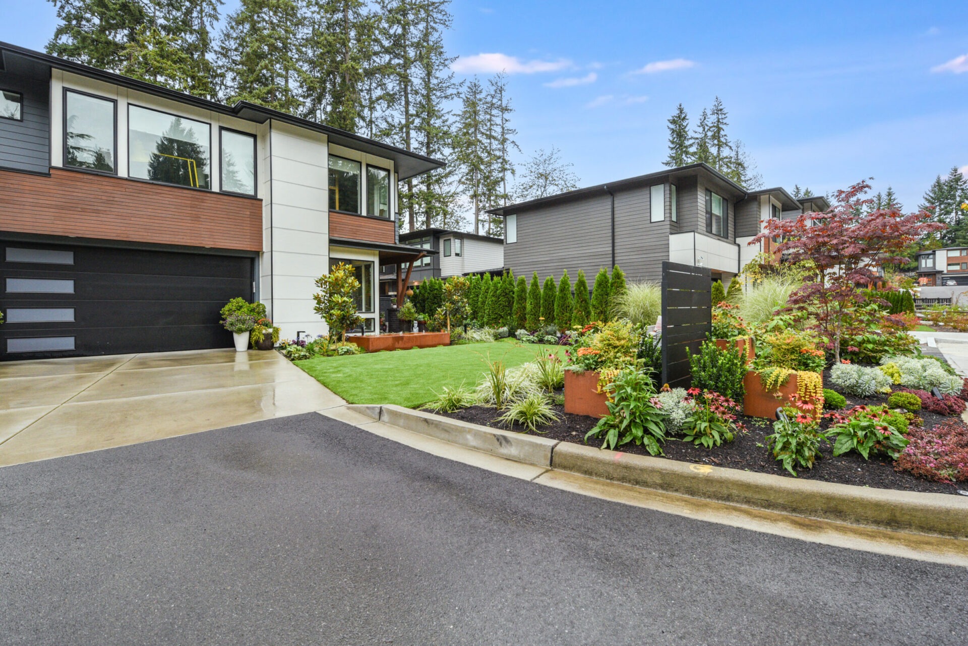 Modern suburban homes with well-maintained gardens on a quiet street. Lush greenery and trees surround the contemporary, minimalist architecture under a clear blue sky.