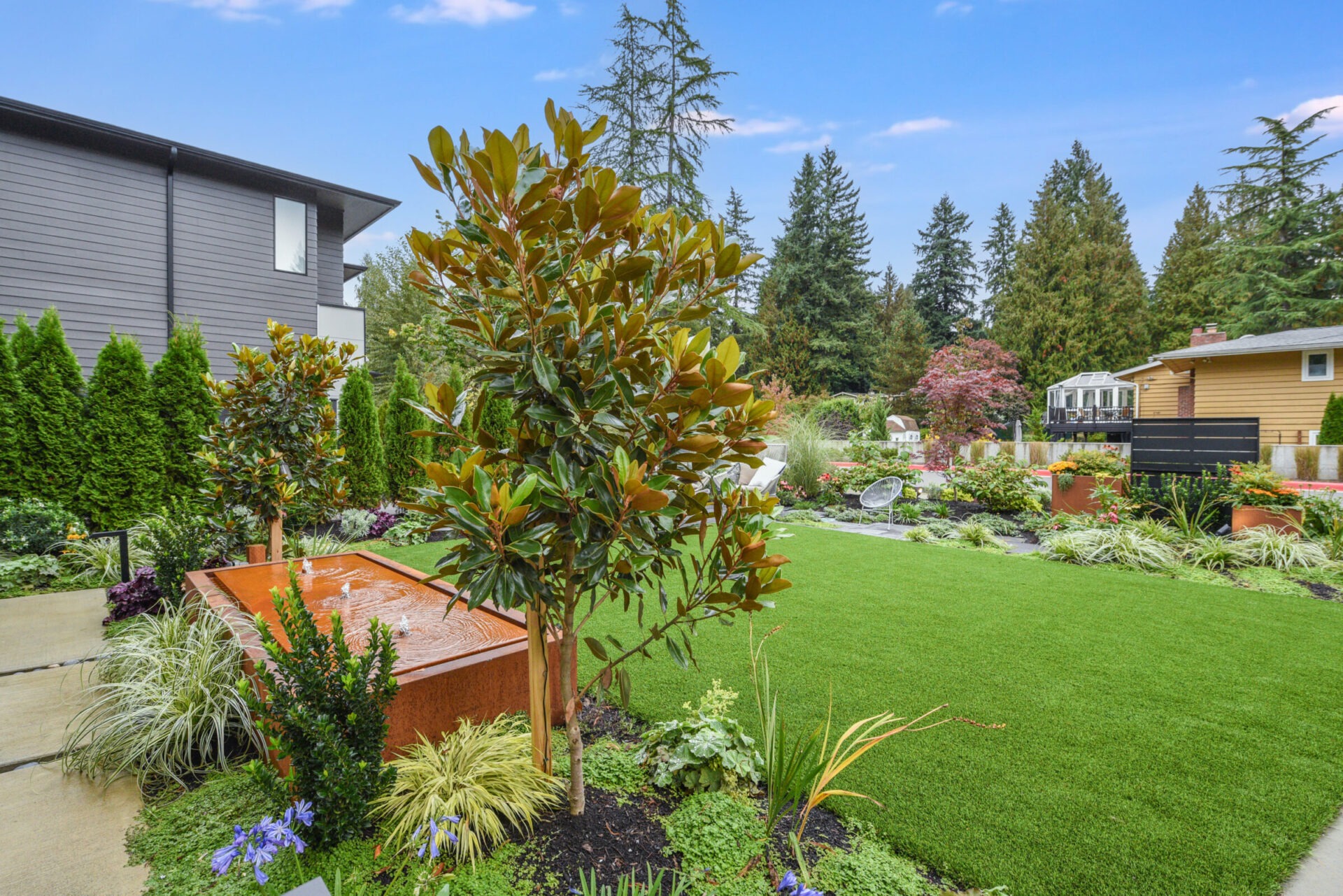 A landscaped backyard features a lush green lawn, diverse plants, and modern decor. Trees and shrubs line the perimeter of the yard.