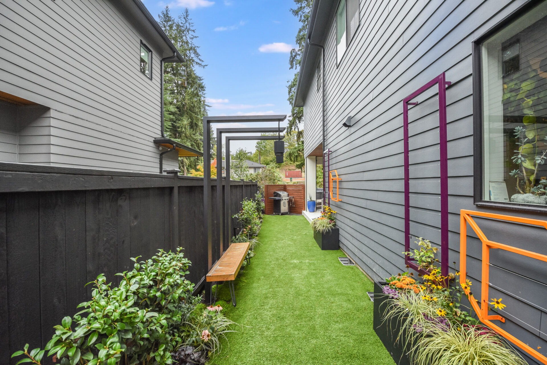 Modern, narrow backyard with artificial grass, bordered by gray buildings and a black fence. Decorative plants and wooden bench enhance the space's cozy feel.