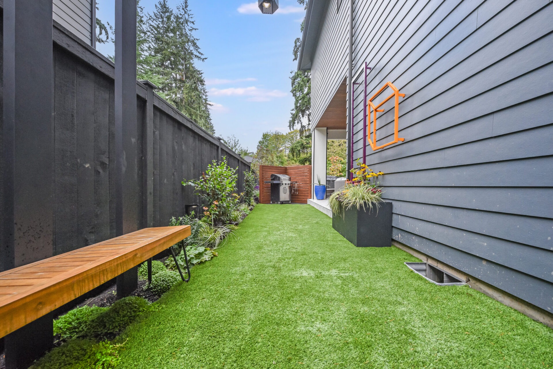 Narrow garden space with artificial grass, wooden bench, flower pots, and a small outdoor grill area. Surrounded by wooden fencing and modern siding.