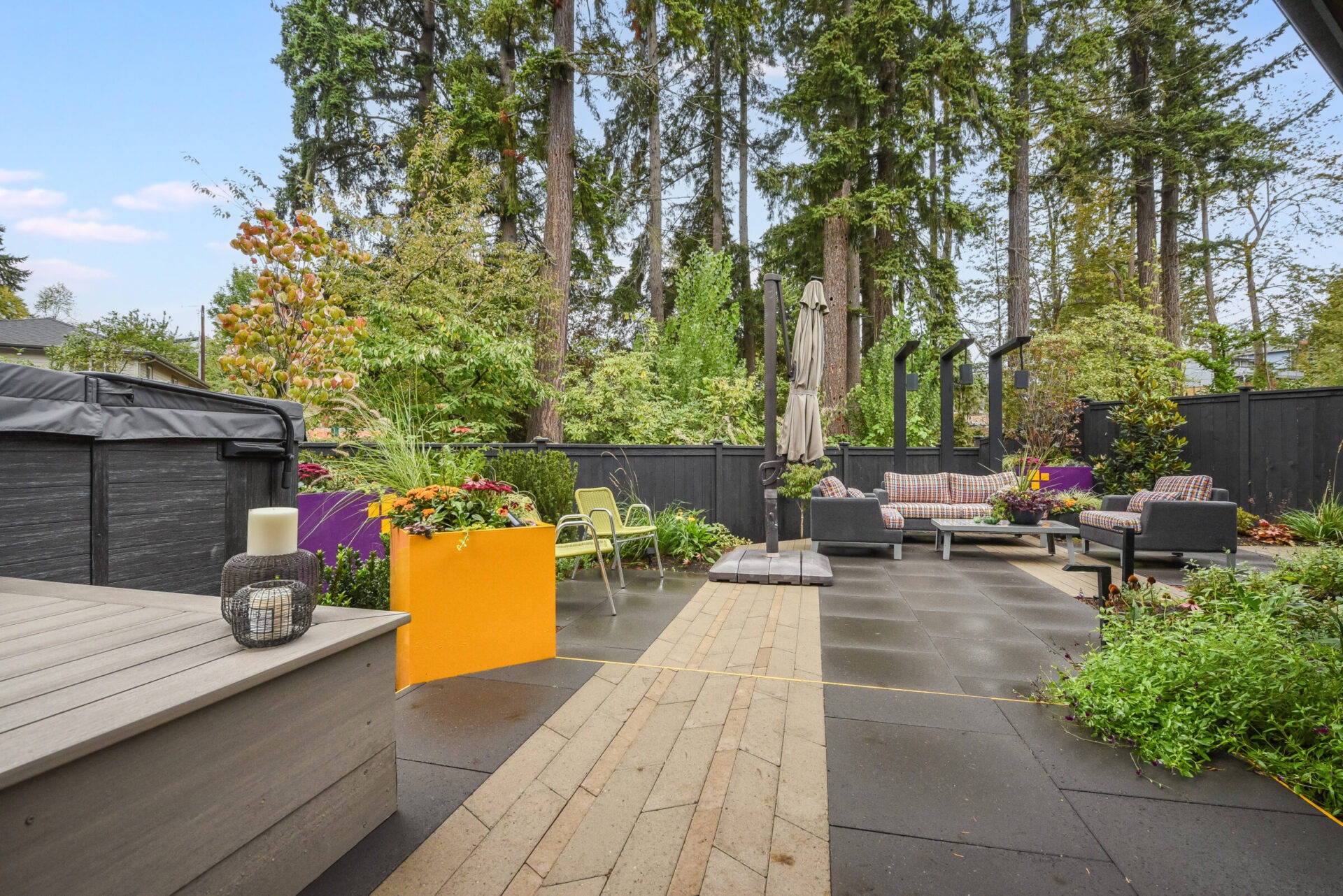 Modern backyard with patio furniture, vibrant planters, and tall trees. Spacious seating area surrounded by lush greenery, creating a serene outdoor retreat.