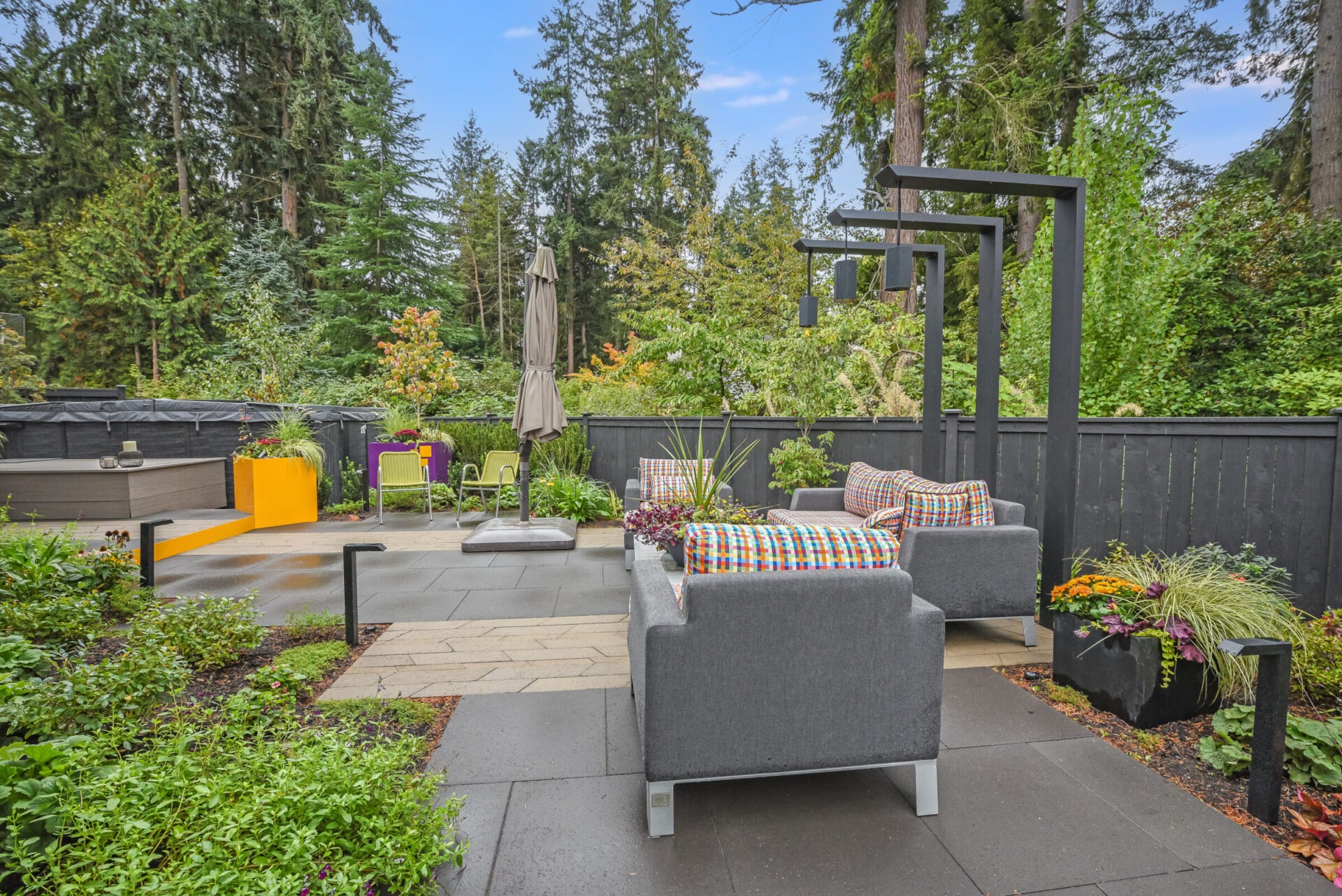 A modern patio with gray furniture, colorful cushions, surrounded by lush greenery and trees, creating a serene outdoor living space.