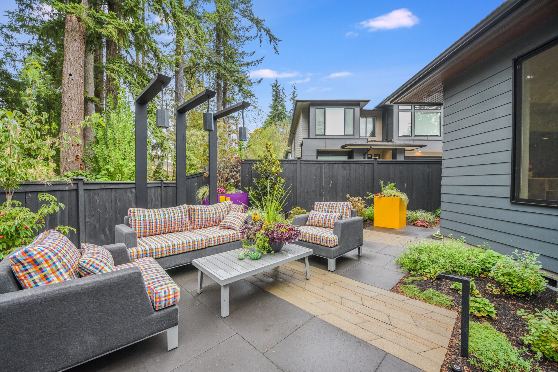 Modern backyard patio with gray furniture, colorful cushions, wood and stone path, surrounded by trees and plants, adjacent to a contemporary house.