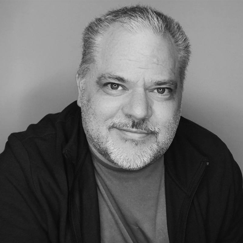 A black and white portrait of a person with short hair and a beard. The person is wearing a dark jacket and a shirt.