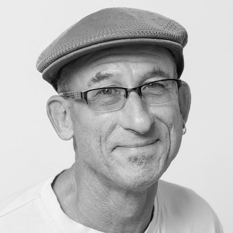 A person wearing a cap and glasses smiles at the camera. The image is in black and white with a plain background.