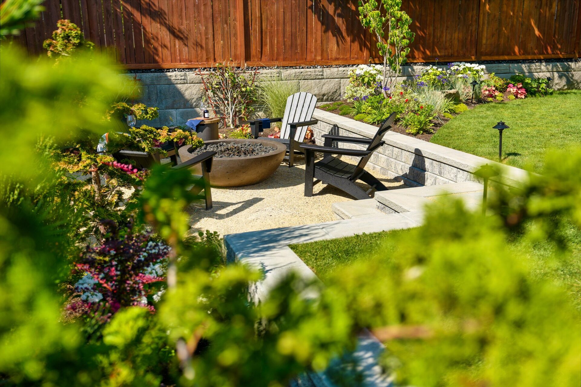 A serene garden patio with Adirondack chairs and a fire pit, surrounded by lush greenery and a wooden fence.