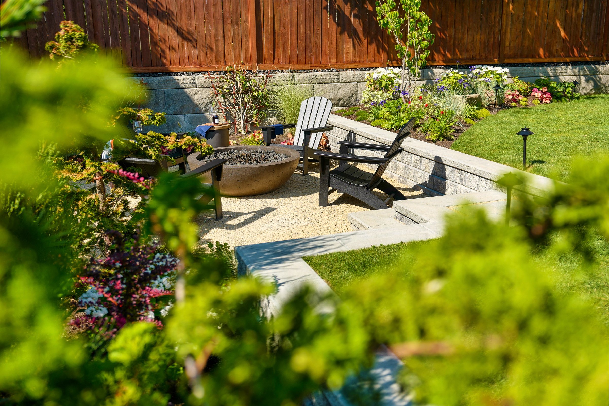 A serene garden patio with Adirondack chairs and a fire pit, surrounded by lush greenery and a wooden fence.