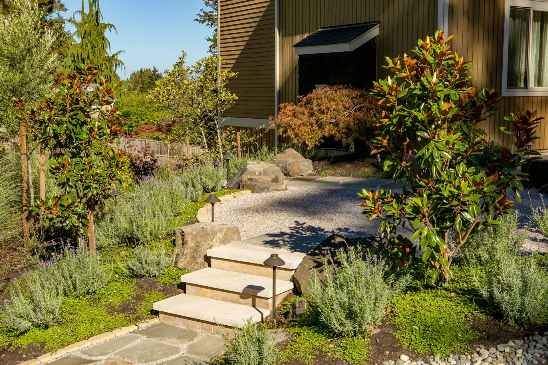 A serene garden pathway with steps leads to a house entrance, surrounded by green foliage and small trees under a clear blue sky.