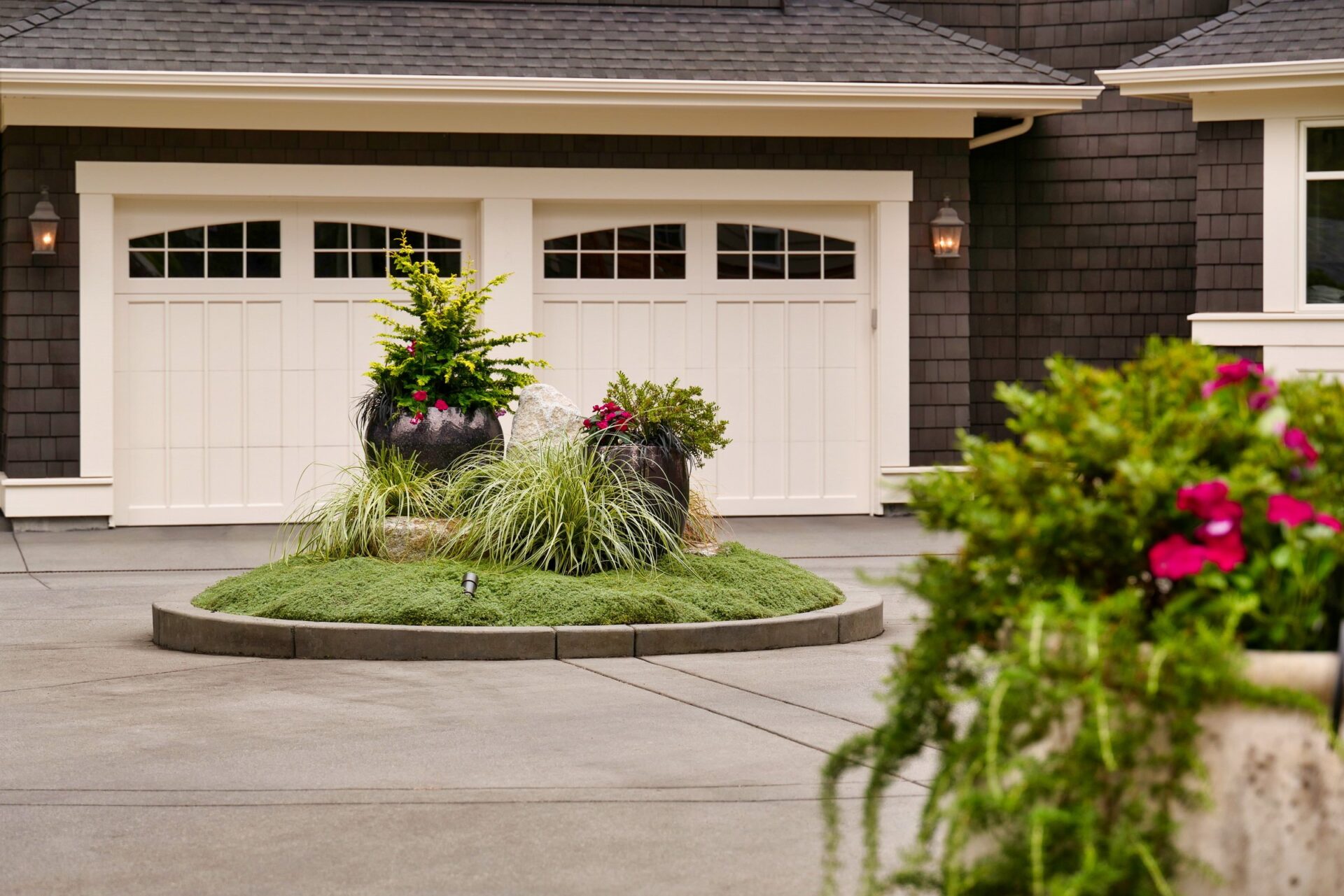 A residential driveway features a rounded garden bed with shrubs and potted plants in front of a double garage with white doors.