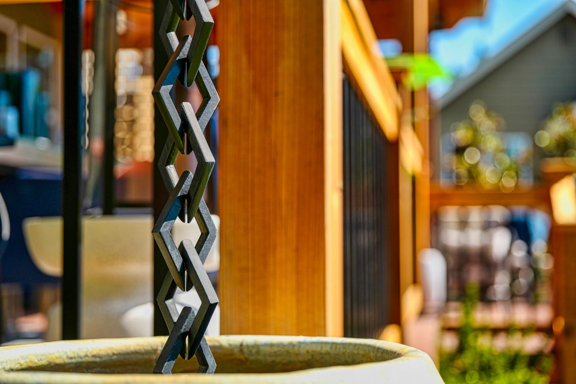 Close-up of a modern metal rain chain with wooden railing and blurred background, featuring a small house and garden under bright sunlight.