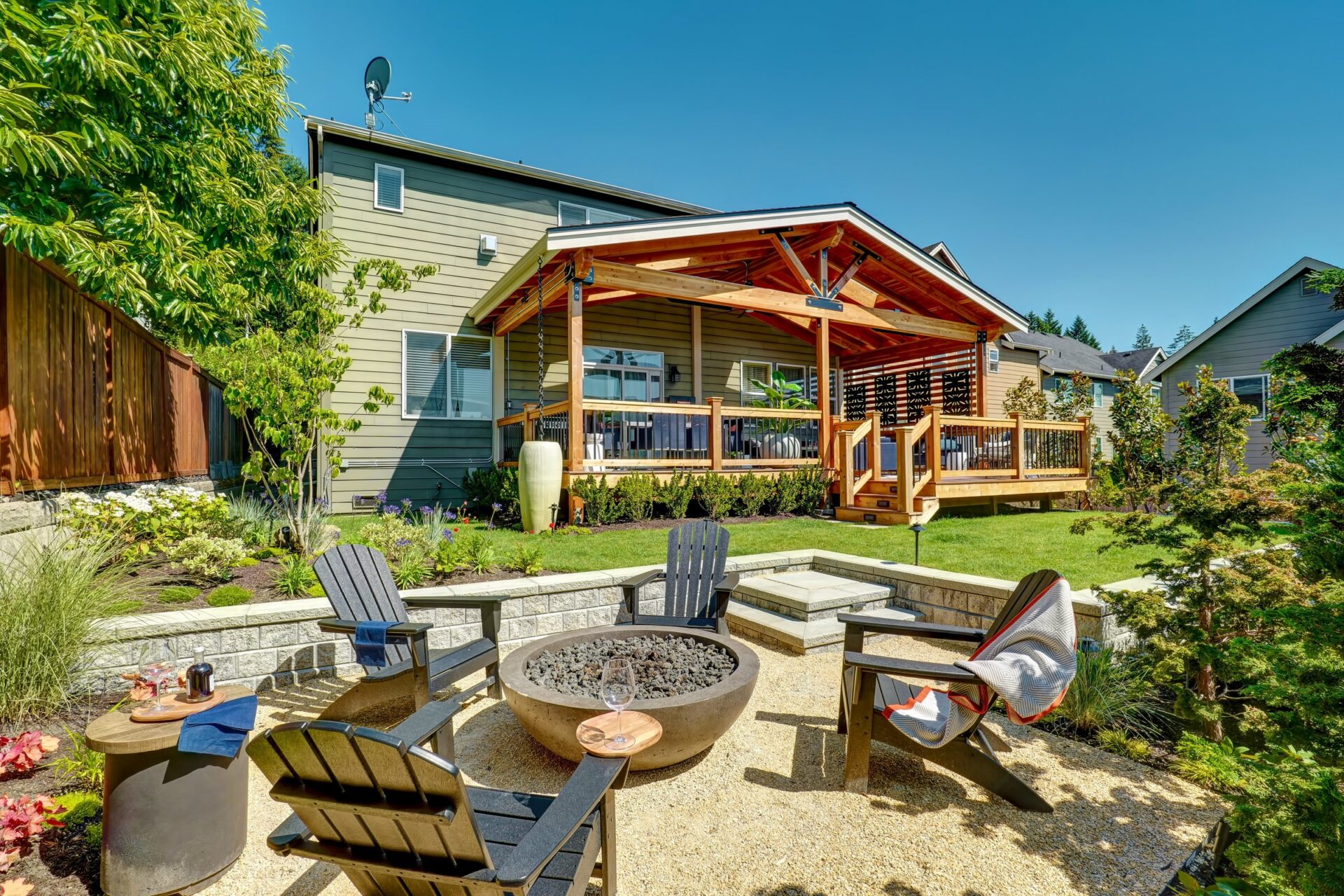 A backyard with a covered patio, fire pit, and wooden chairs. Surrounded by lush greenery, the area is ideal for relaxing outdoors.