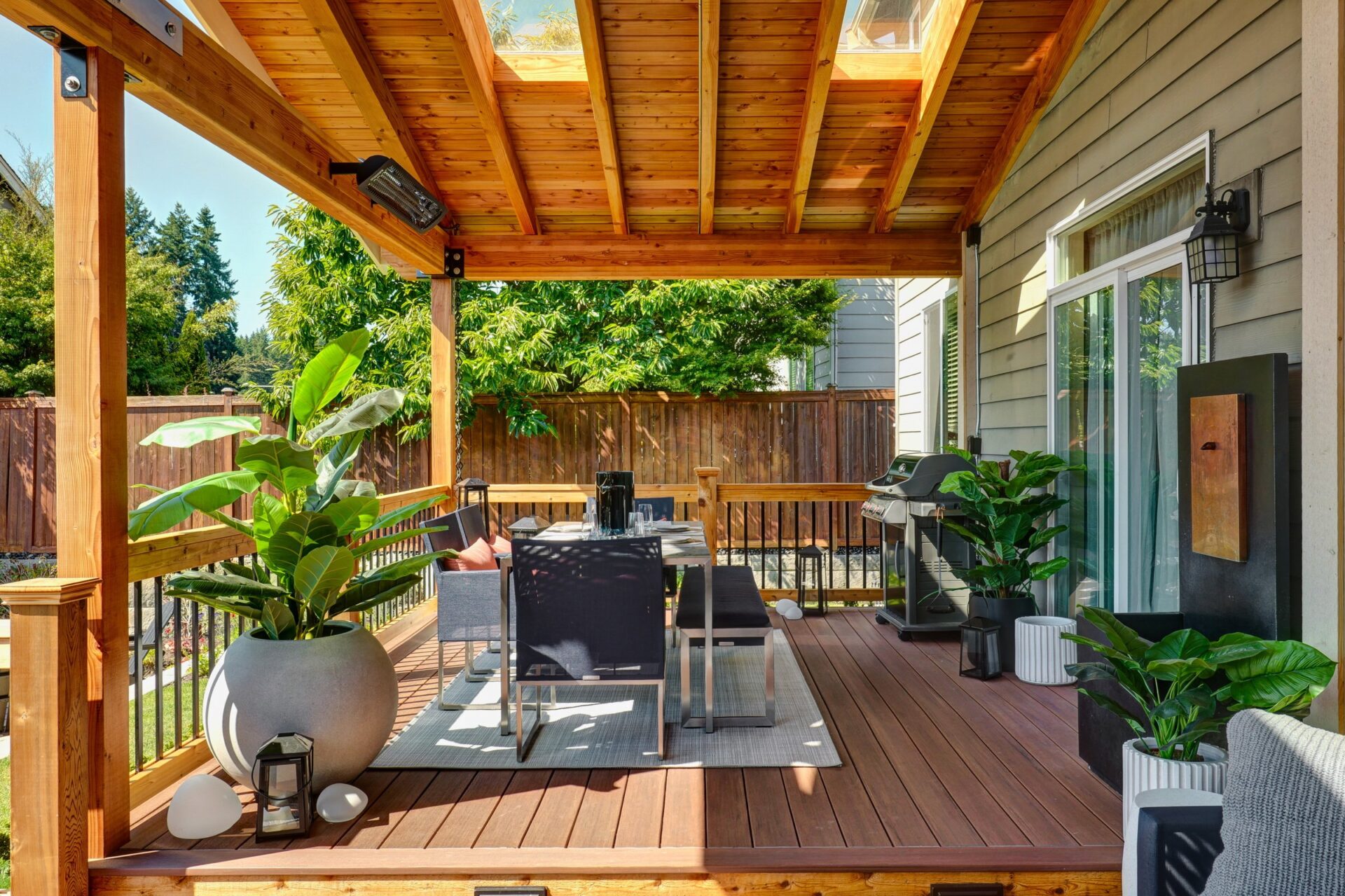 A cozy wooden deck with outdoor seating, potted plants, and a grill. The area is sheltered by a slatted roof, overlooking a fenced yard.