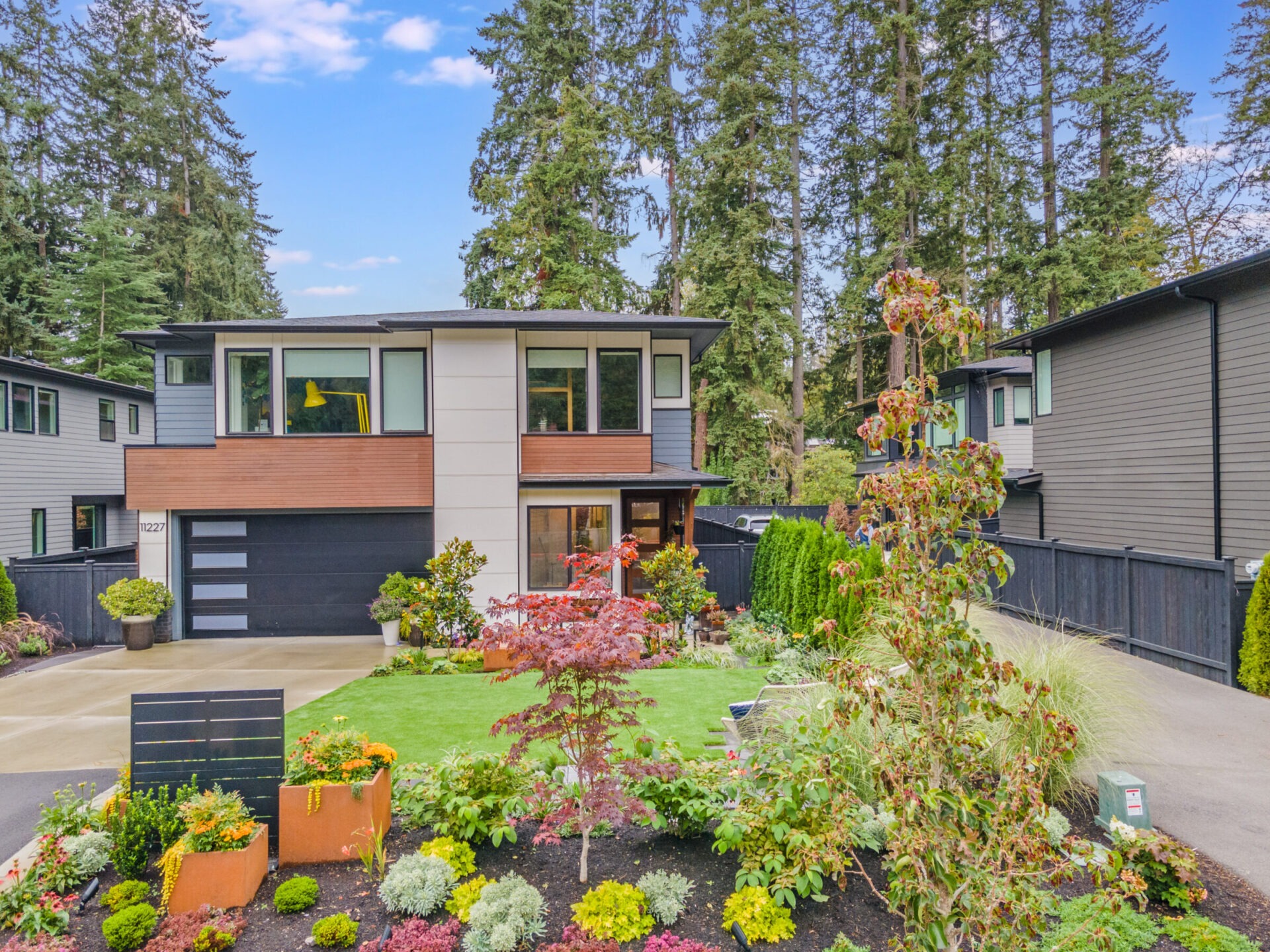 Modern two-story house surrounded by lush greenery and landscaped garden. Trees in the background, clear sky. No people or landmarks visible.