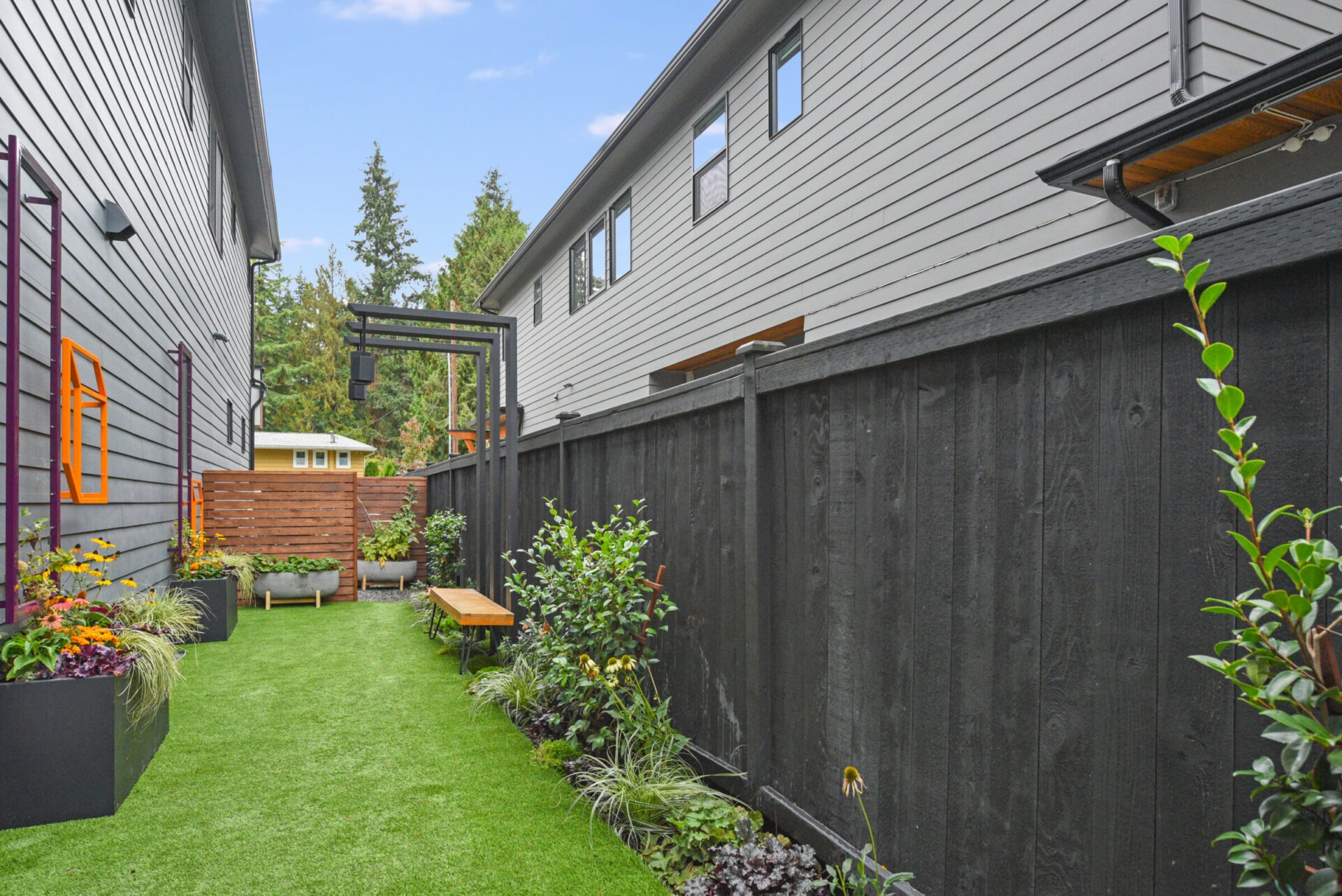 Narrow backyard with artificial grass, black fence, flanked by two gray houses. Potted plants and small bench enhance the cozy atmosphere.