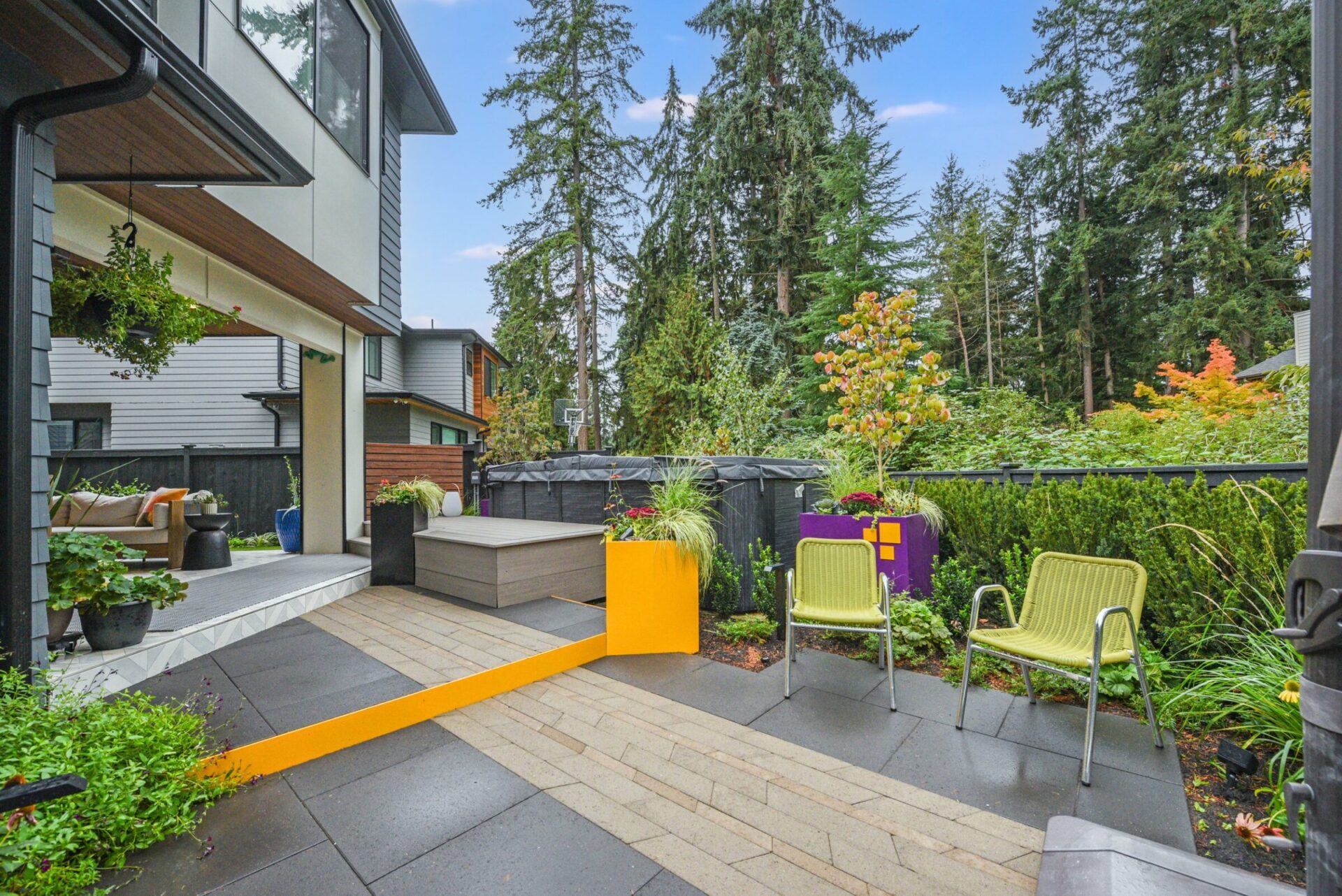 Modern patio with colorful chairs, surrounded by lush greenery and tall trees. Contemporary house exterior features large windows and clean lines, creating a tranquil atmosphere.