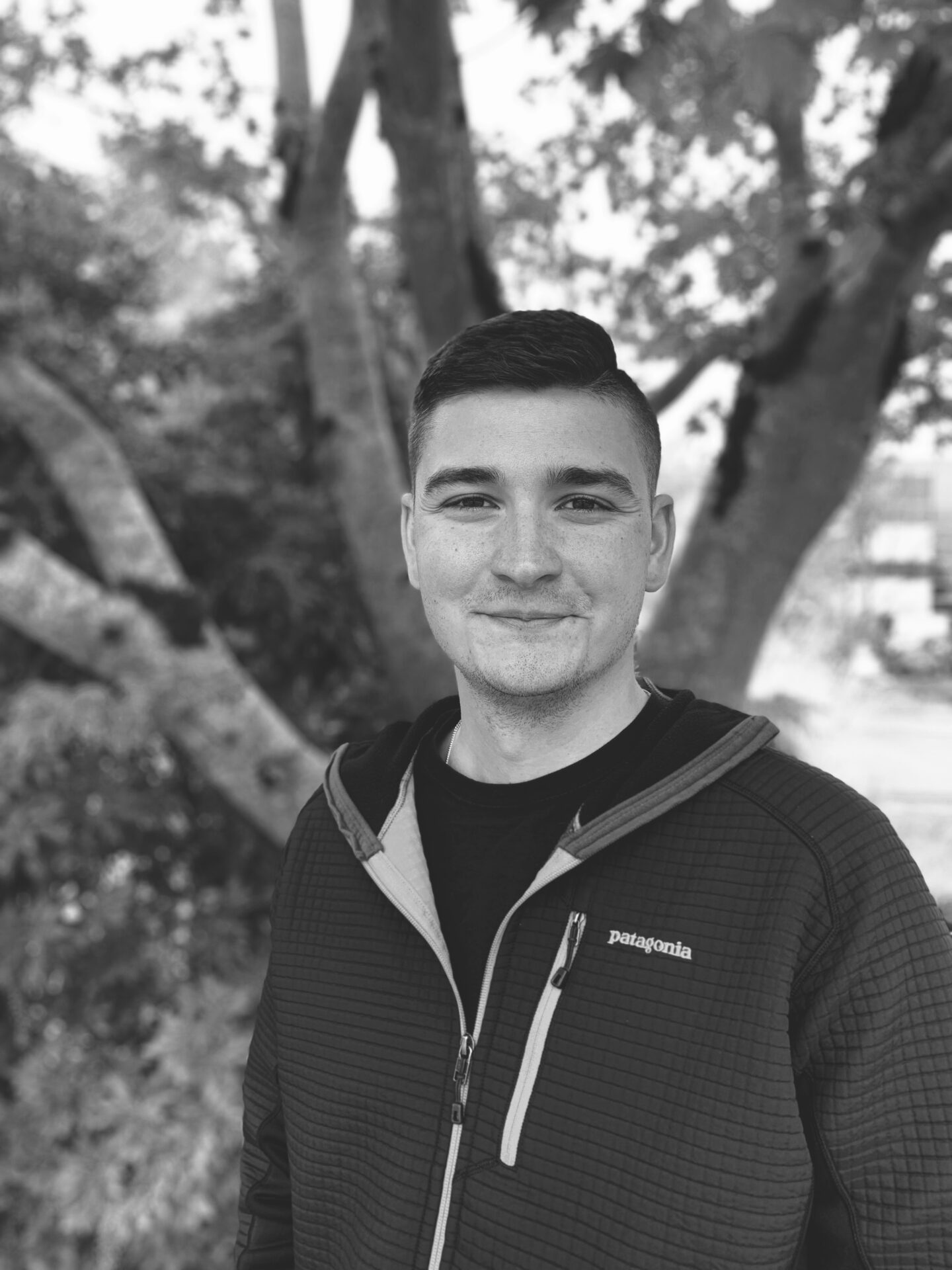 A person stands outdoors in front of a tree, wearing a jacket, smiling, in a black and white portrait. No recognizable landmarks visible.